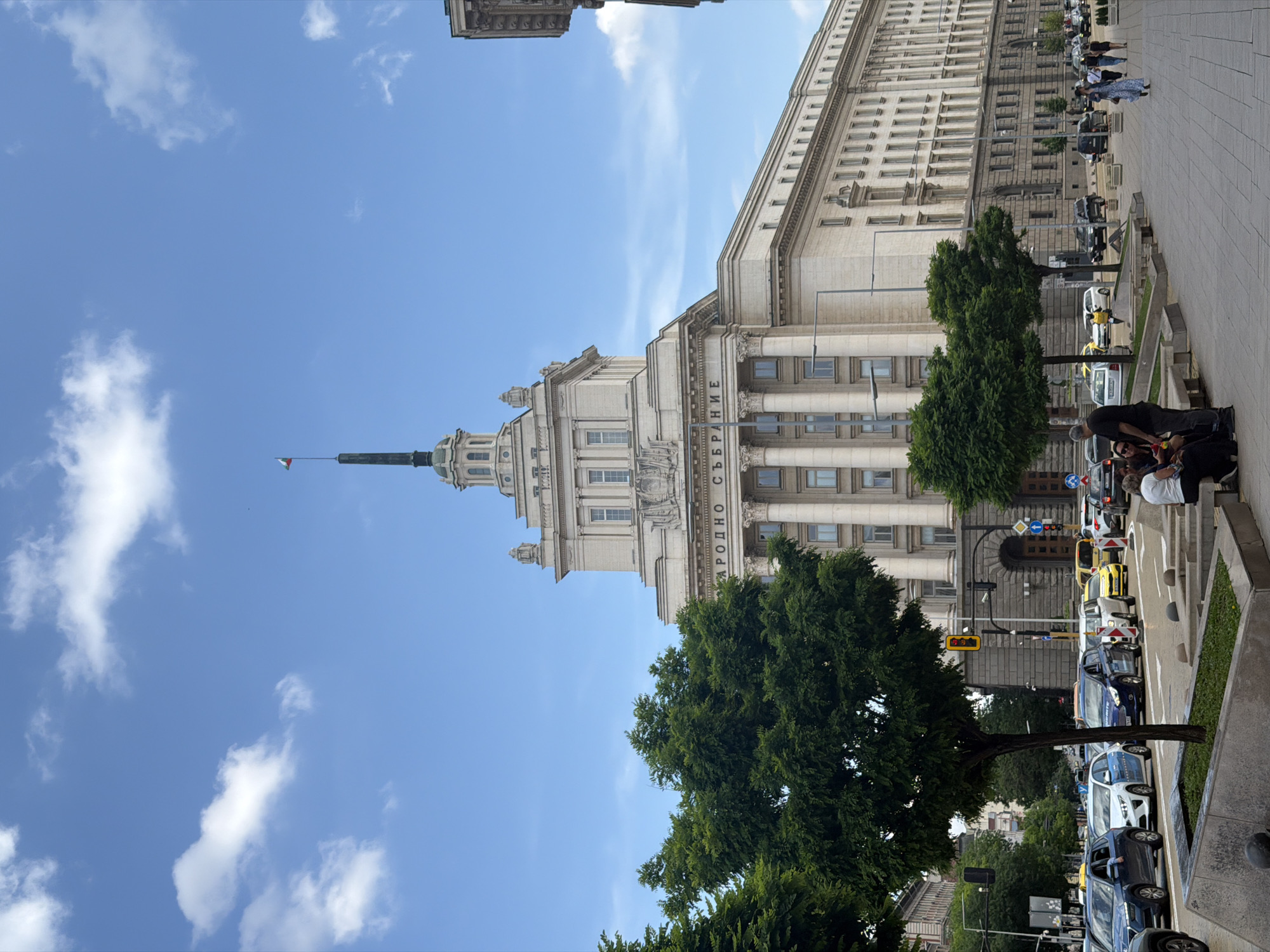 A bright daytime street scene in Sofia, Bulgaria, features a grand, imposing building under a clear blue sky with scattered white clouds. The main subject is a large, light-colored building with classical architectural elements, including prominent columns and an ornate facade. A tall spire with a small Bulgarian flag (white, green, red) at its peak rises from the center of this building. On the building's facade, Cyrillic text is visible, reading "НАРОДНО СЪБРАНИЕ" (Narodno Sabranie), which translates to "National Assembly" or "People's Assembly," identifying it as the Bulgarian Parliament.
In the foreground and to the right, a busy city street runs parallel to the building. Numerous cars, including several yellow taxis, are parked or moving slowly. A wide sidewalk with paved stones separates the street from the building, where several people are seen walking or standing. In the lower right, a group of people is visible, with one person in a white shirt and another in a dark shirt appearing to be interacting with something or someone close to the ground. Lush green trees line parts of the sidewalk and street, providing shade. A yellow traffic signal with a numerical display is also visible near the road. The scene depicts typical urban activity on a sunny day.