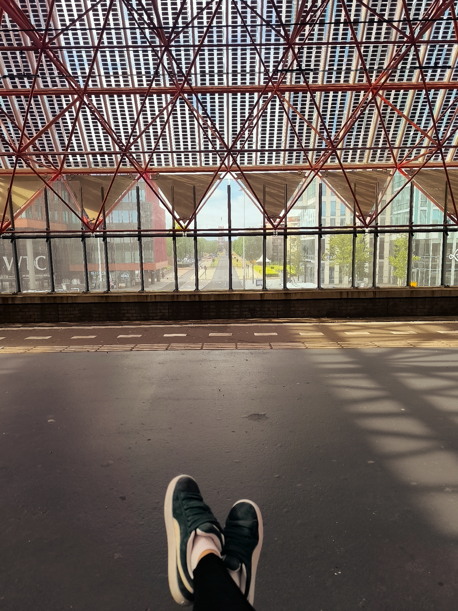 The image shows a person's legs and feet wearing dark sneakers with white soles and laces, and white socks, as they sit on a dark gray asphalt surface. The person is facing away from the camera. In the background, there is a train station platform with tactile paving and a long horizontal row of windows. Beyond the windows, a street with buildings, trees, and a yellow vehicle can be seen. Above the platform, there is a distinctive red geometric roof structure with numerous rectangular solar panels. This is the characteristic roof of Almere Central Station in Almere, Netherlands. The scene is illuminated by natural light, casting shadows from the roof structure onto the platform.