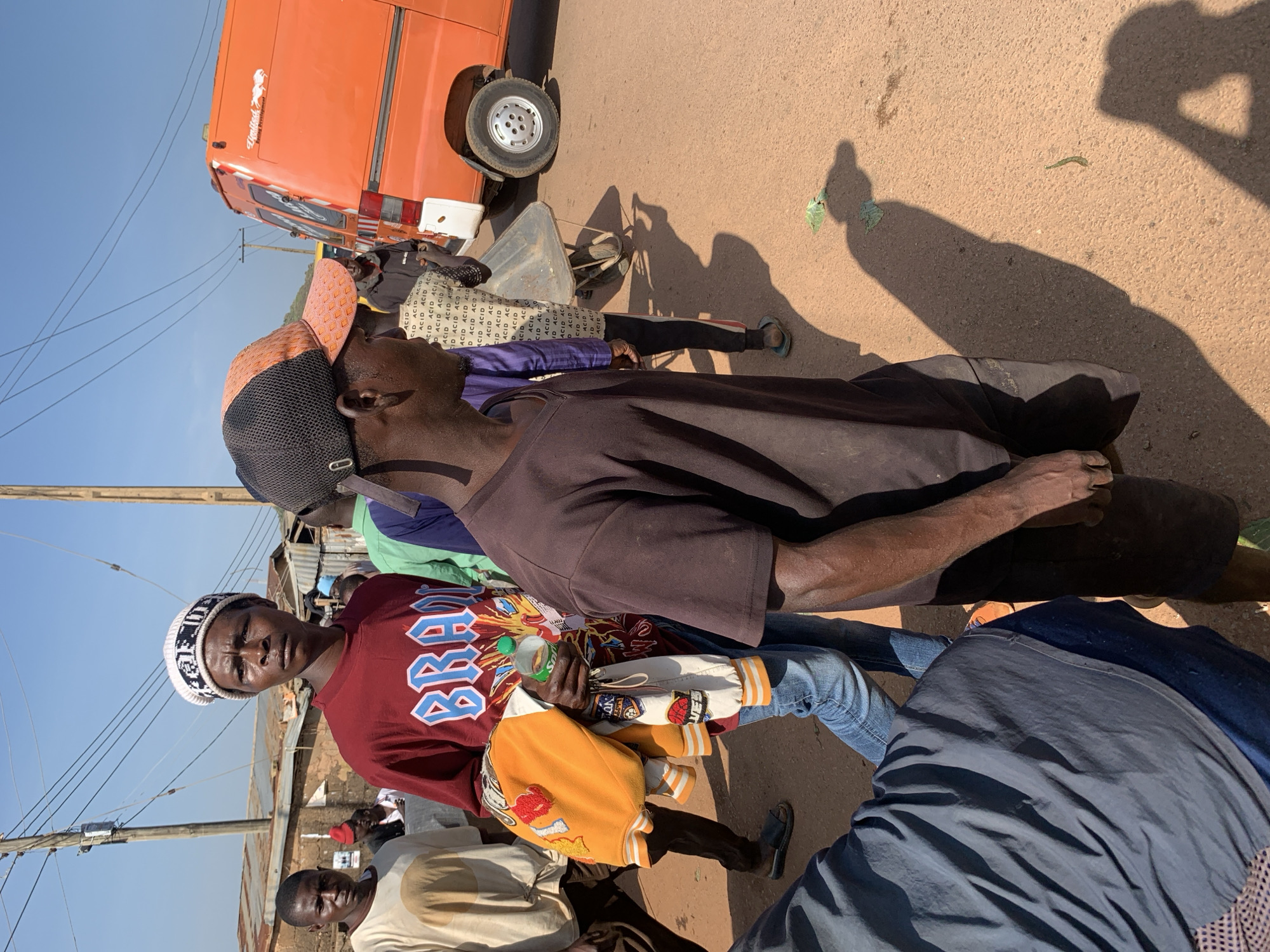 In Gura Top, Nigeria, under a clear sky, a group of men walk along a dirt road lined with simple buildings. The foreground features a man in a dark brown t-shirt and shorts, his gaze directed forward and to the right. He is shadowed by another person in a dark blue garment, whose lower body is visible. To the left, a man in a maroon t-shirt with the word "BRAVE" emblazoned in blue letters and a white beanie walks with a yellow and white jacket draped over his arm, holding a small green bottle. Behind him, a man in a light-colored t-shirt stands. Further back, an orange van is parked on the right side of the road. In the middle ground, a man is pushing a wheelbarrow. The overall scene depicts a slice of daily life in a Nigerian town.
