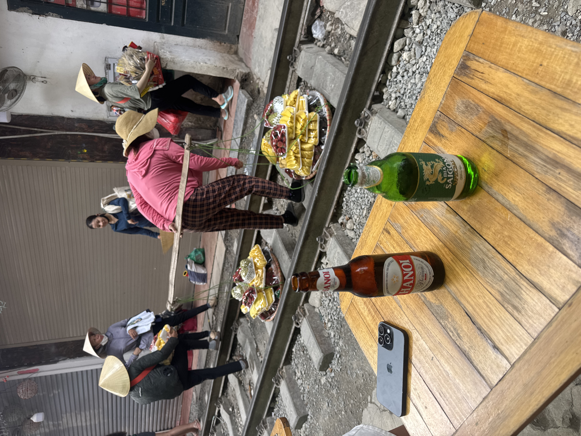 The image captures a dynamic street scene on the famous Hanoi Train Street in Hà Nội, Vietnam. In the foreground, a light brown wooden table displays a dark gray smartphone with a triple-lens camera array and two beer bottles: a brown bottle of "Hanoi Premium" beer and a green bottle of "Bia Saigon Special."
Beyond the table, the central focus is on a set of active railway tracks, consisting of steel rails mounted on concrete sleepers, running through a narrow urban corridor. Several individuals are present on or immediately beside the tracks. A woman in a pink long-sleeved top and plaid pants is seen bent over, utilizing a wooden shoulder pole (quang gánh) to carry two large, round woven baskets. These baskets, positioned between the rails, are filled with numerous brightly colored, individually plastic-wrapped items, appearing to be various fruits or snacks (such as yellow mango slices, red strawberries, and green items).
Further down the tracks, another woman wearing a light grey hooded top and a traditional conical hat (nón lá) stands holding a white cloth item and a bag. To her right, a third woman, also in a conical hat and a green short-sleeved top, carries a red plastic basket overflowing with numerous packaged goods. In the background, various urban structures line the tracks, including buildings with a large grey metal roller shutter and a white wall featuring a mounted electric fan. Other individuals and various bags or containers are also visible in the periphery. The ground around the tracks is composed of gravel and dirt.