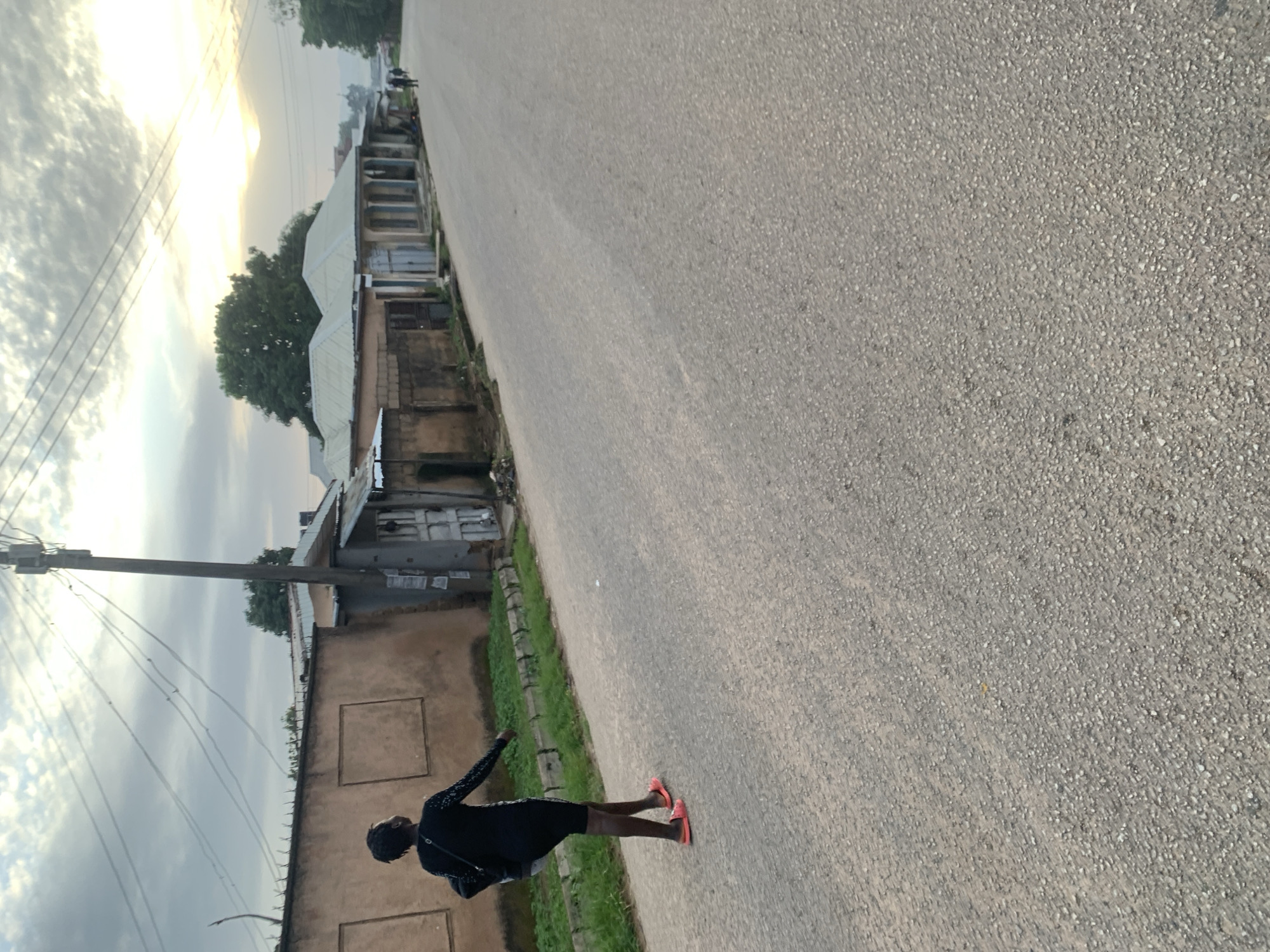 A full shot captures a young woman walking away from the camera on a paved road in Jos, Nigeria. The woman is wearing a black, long-sleeved dress with white detailing on the sleeves and is carrying a black bag over her left shoulder. She has on bright orange sandals. The road is lined with buildings on the left side, which appear to be residential or small shops, with corrugated metal roofs and light-colored walls. Some of the buildings have a weathered appearance. There is a patch of green grass and some weeds along the side of the road in front of the buildings. A utility pole with multiple wires stretches across the frame above the buildings. The sky is cloudy with a hint of sunlight breaking through in the distance. Two figures can be seen further down the road in the background, walking away from the camera. The overall scene suggests a typical street in a Nigerian city.