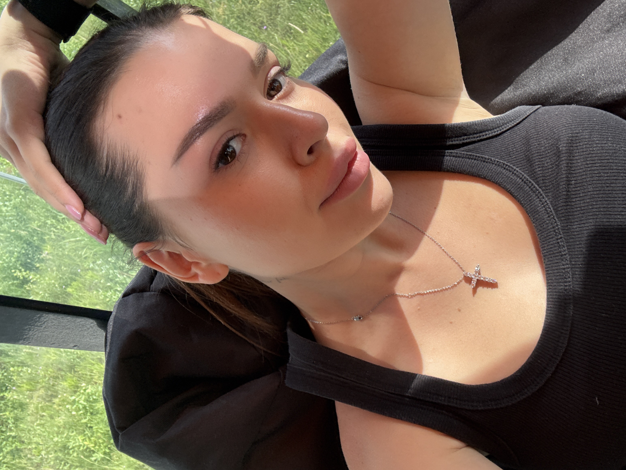 A medium close-up, eye-level selfie of a young woman with dark hair pulled back, taken outdoors. She is wearing a black ribbed tank top and a silver necklace with a cross pendant. Her right hand is on top of her head. Sunlight casts a warm glow on her face and chest. The background is blurred, showing green foliage and a dark object, possibly a chair. There is no indication of Prague, Czechia in the image.