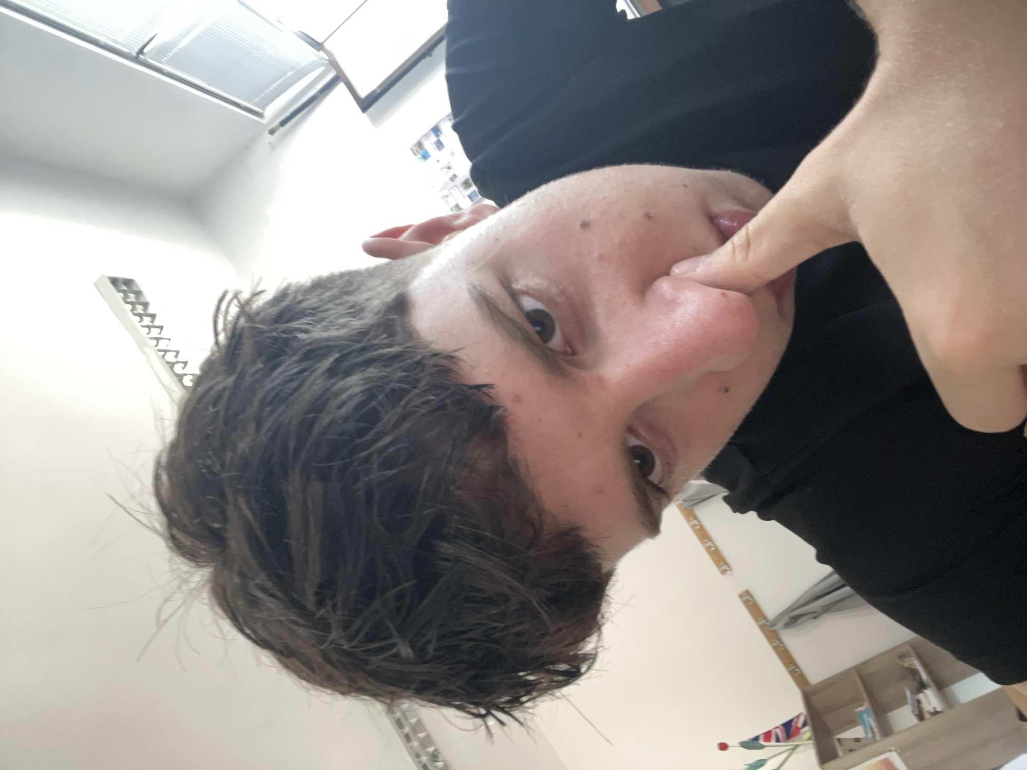 An indoor, eye-level shot captures a young person, likely male, with their head tilted and eyes looking directly at the camera. The image is rotated on its side, presenting a unique perspective. The person's left eye is prominent, with dark brown hair appearing slightly wet or messy across their forehead and to the left. Their skin tone is fair, with some small blemishes visible on the forehead and cheek. An index finger of their right hand is playfully inserted into their mouth, touching the inner lip or tongue area. They are wearing a black top or shirt, visible on their right shoulder and arm.
The background reveals a light-colored room, possibly in Zlatograd, Bulgaria. Overhead, a large rectangular light fixture with louvers is visible, suggesting artificial lighting. Another, simpler white rectangular fixture is on the left wall. Towards the bottom right, parts of a wooden shelving unit or desk are discernible, holding various indistinct objects. A small portion of a Union Jack flag and what appears to be a decorative red flower are also visible in the lower right corner, adding a touch of color to the otherwise neutral background. The overall lighting is bright, illuminating the scene clearly. No discernible text is visible in the image.