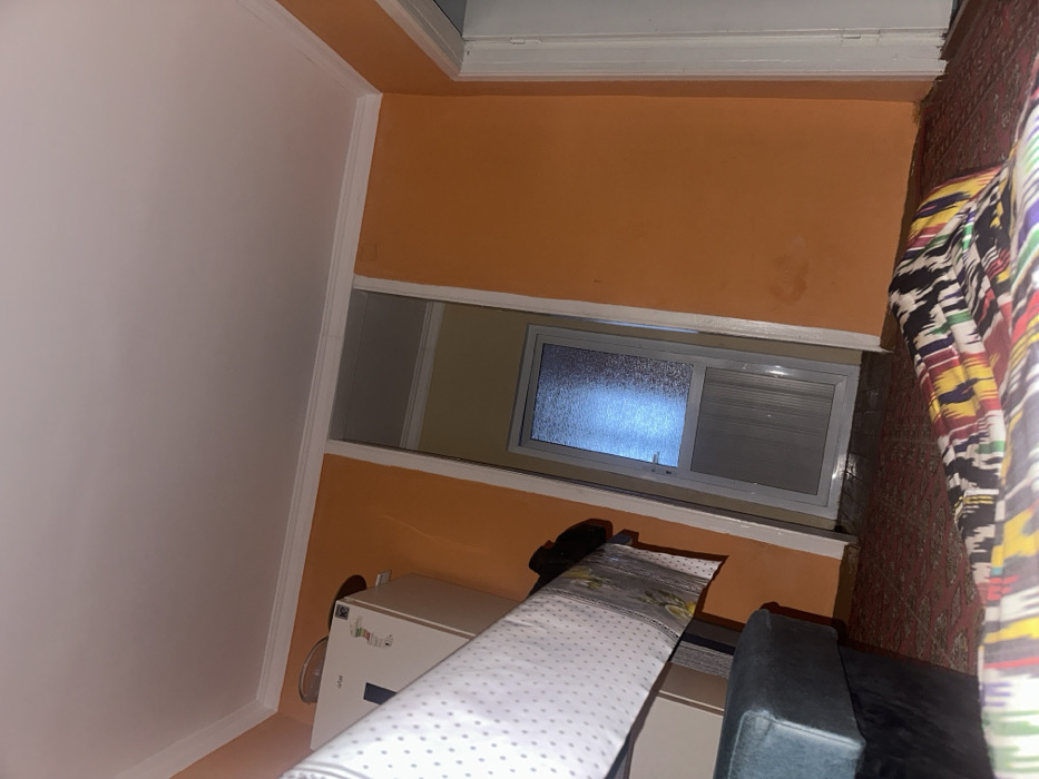 A sideways view of a room with orange walls and white trim. A window with frosted glass sits between two sections of wall. Below the window is a white appliance, possibly a refrigerator or freezer. In the foreground, a bed with a white polka dot cover and a green chair are partially visible. A colorful curtain hangs to the right side of the room.