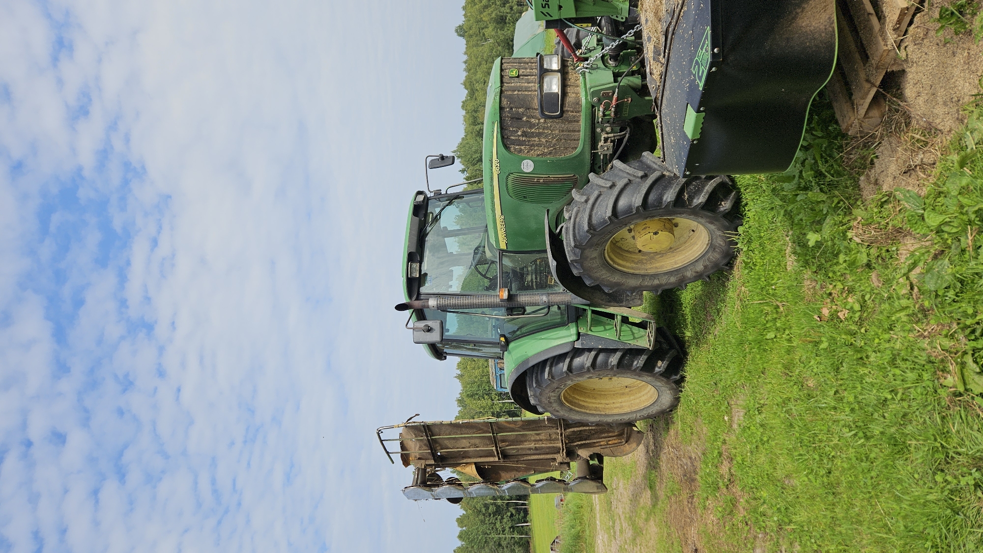 A green John Deere tractor is parked in a grassy field in Kapūņi, Latvia. The tractor has large, black tires with yellow rims. Attached to the rear of the tractor is a large, metallic piece of equipment, possibly a harvester or a baler. In the foreground, a black cylindrical object with green accents is placed on a wooden pallet. The background features a dense line of green trees and a partly cloudy blue sky. The scene appears to be set in a rural or agricultural area.