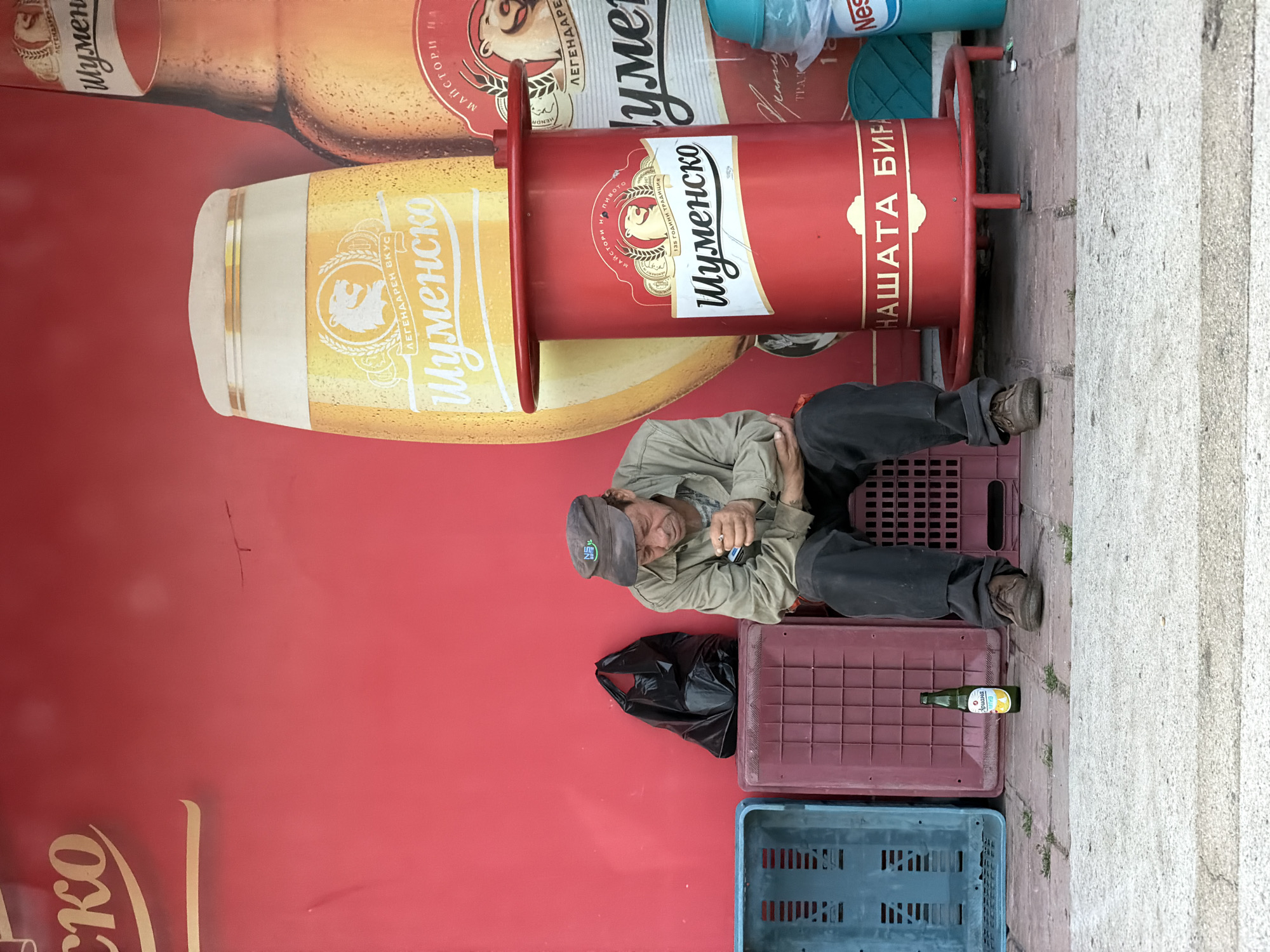 The image depicts a man lying down on plastic crates near a large advertisement for "Shumensko" beer in Burgas, Bulgaria. The man is wearing dark pants, a light-colored jacket, and a cap. He appears to be holding something in his hands. A black plastic bag and an empty bottle are near him. The background includes the advertisement for "Shumensko" beer, a red cylindrical structure with "Shumensko" branding, and part of a blue container labeled "Nestle". The setting seems to be an outdoor public space with concrete pavement and steps visible on the right side of the image.