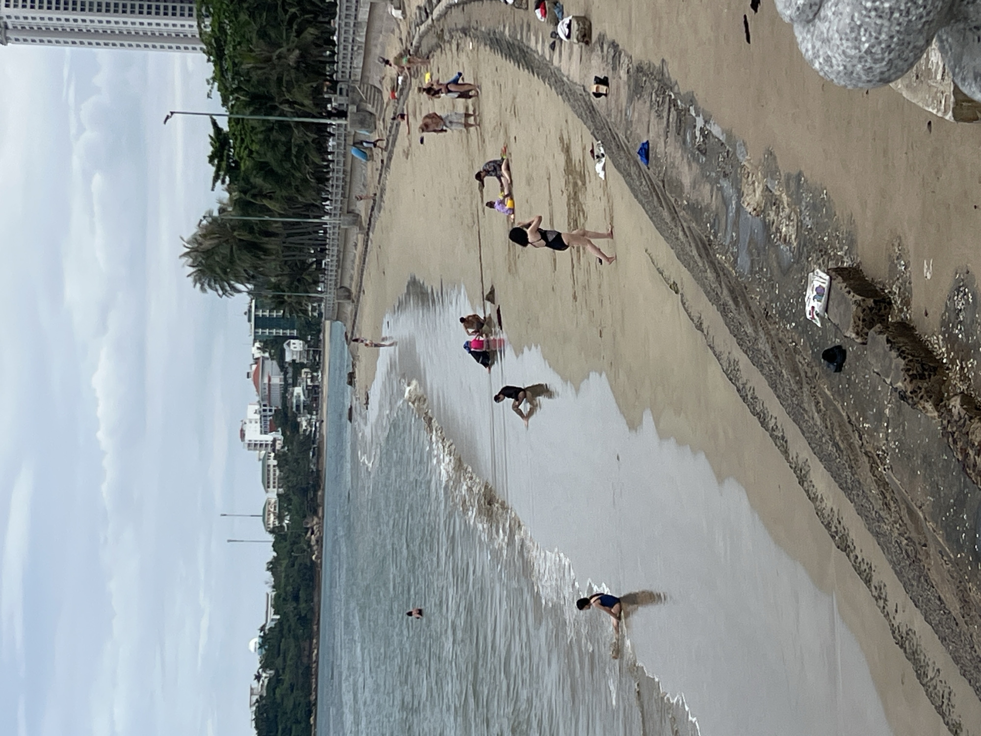 The image depicts a lively beach scene in Bac Nha Trang Ward, Vietnam, under an overcast sky. A wide sandy beach stretches along a calm body of water, likely the ocean, with gentle waves breaking near the shore. A concrete promenade or barrier separates the beach from a verdant strip of palm trees and urban architecture.
Numerous individuals are present on the beach and in the shallow water. People are engaged in leisurely activities such as wading, walking along the water's edge, and relaxing on the sand. Some are seen bending down at the waterline, possibly interacting with the water or sand. Various personal items, including bags and clothes, are scattered on the concrete barrier.
In the background, a dense line of palm trees borders the coastline, behind which several buildings are visible, including a tall high-rise structure and other multi-story buildings. Two streetlights are also present along the promenade. The sky is covered with light clouds, indicating an overcast day or a time of day with indirect sunlight. A white wrapper with illegible text is visible on the sand near the concrete barrier. The overall atmosphere appears relaxed, with people enjoying the coastal environment.