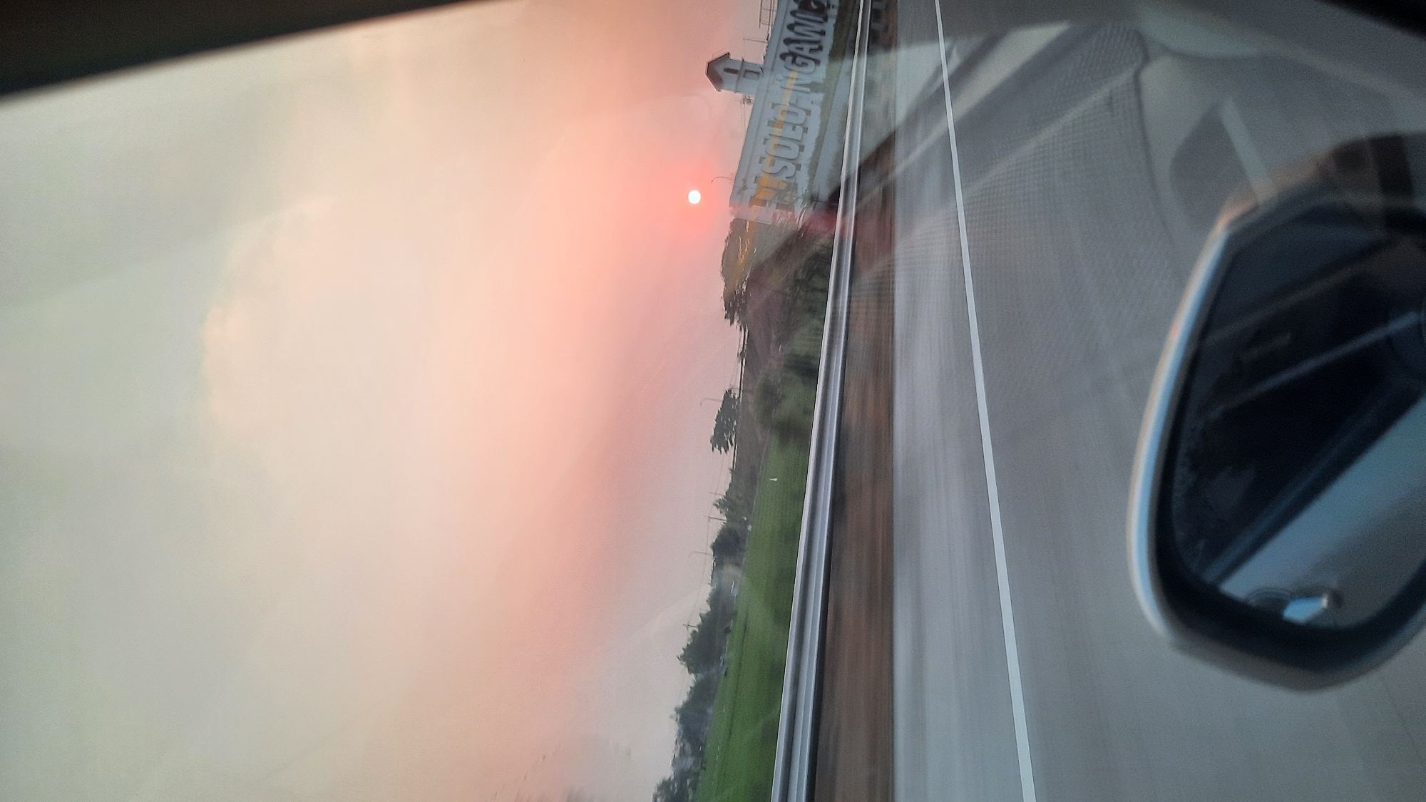 The image is a photograph taken from inside a vehicle, likely in motion, looking out towards a sunset. The sky is filled with a warm gradient of pink and orange, indicating either sunrise or sunset. In the distance, there is a bright sun visible, appearing as a small, intense orb.
To the left of the sun, a structure with the text "SOLO-NGAWI" is visible, suggesting the location is near or on the road between Solo and Ngawi, Indonesia. The "SOLO-NGAWI" text is part of a white building or sign. The landscape beyond the road includes green fields and trees, indicating a rural or semi-rural setting. A highway guardrail runs horizontally across the middle ground of the image.
In the foreground, the side mirror of the vehicle is partially visible on the bottom left, reflecting the interior of the car. The road itself, with a white line marking a lane, is also prominently featured. The overall impression is a scenic drive during a colorful twilight.