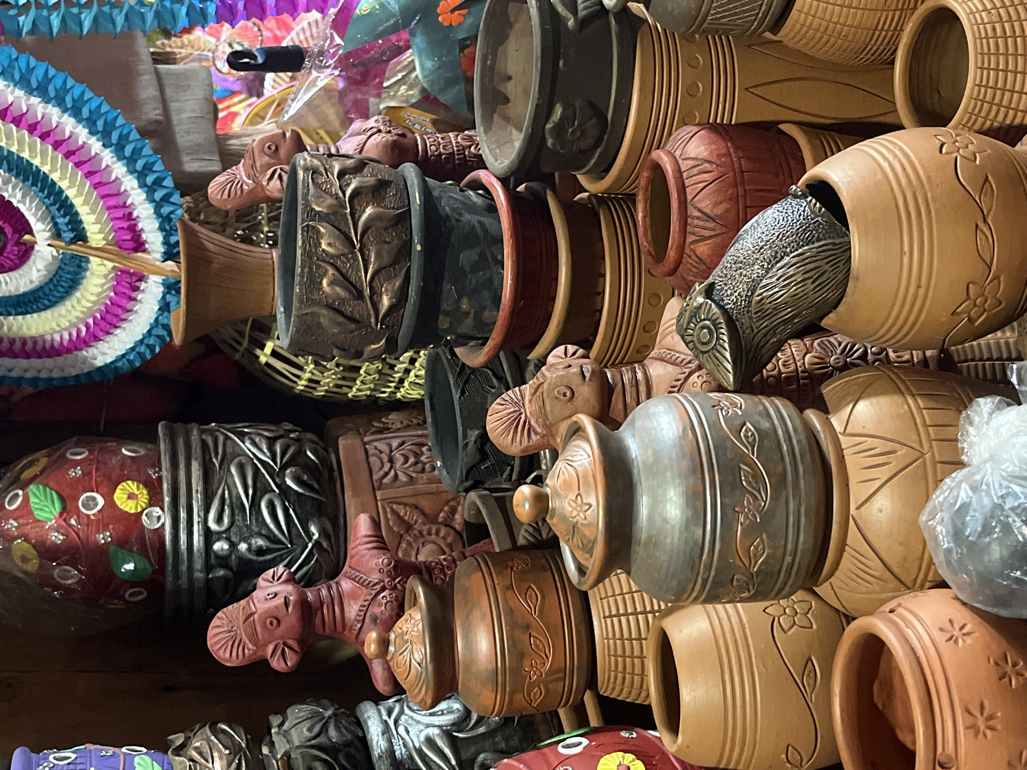 The image shows a collection of earthenware pots and figurines, likely for sale in a market or shop. The pots vary in size, shape, and color, ranging from light terracotta to dark brown and black. Many are decorated with incised or applied designs of flowers, leaves, and geometric patterns. Some of the pots are stacked inside others. There are also small, terracotta figurines of animals, possibly elephants or cows, interspersed among the pots. In the background, there are colorful decorations, including a paper fan and garlands. Based on the style of the pottery and decorative items, the location is likely in Dhaka, Bangladesh. The image depicts a cluttered, artisanal scene.