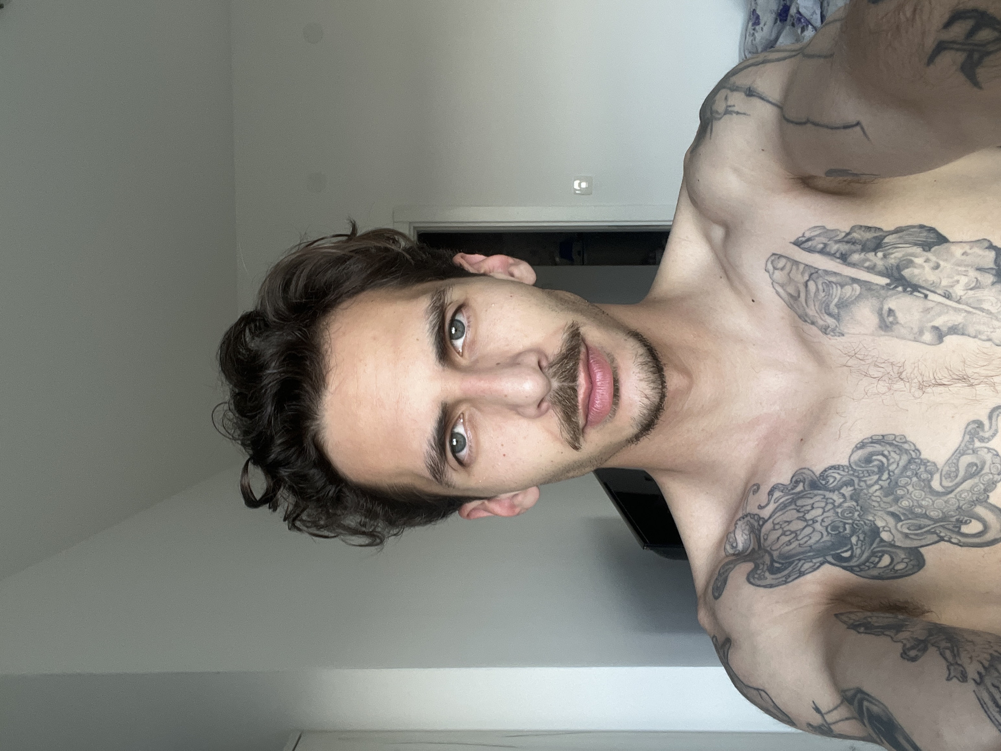 A light-skinned man with dark brown, curly hair, hazel eyes, and a mustache and goatee is depicted from the chest up. The man is shirtless and has multiple tattoos on his chest and shoulders, including a figure of a god, and an octopus. The background is a plain, light-colored wall, and a door frame is visible behind the man's head. There is nothing in the image to indicate the location of Rafailovići, Montenegro.