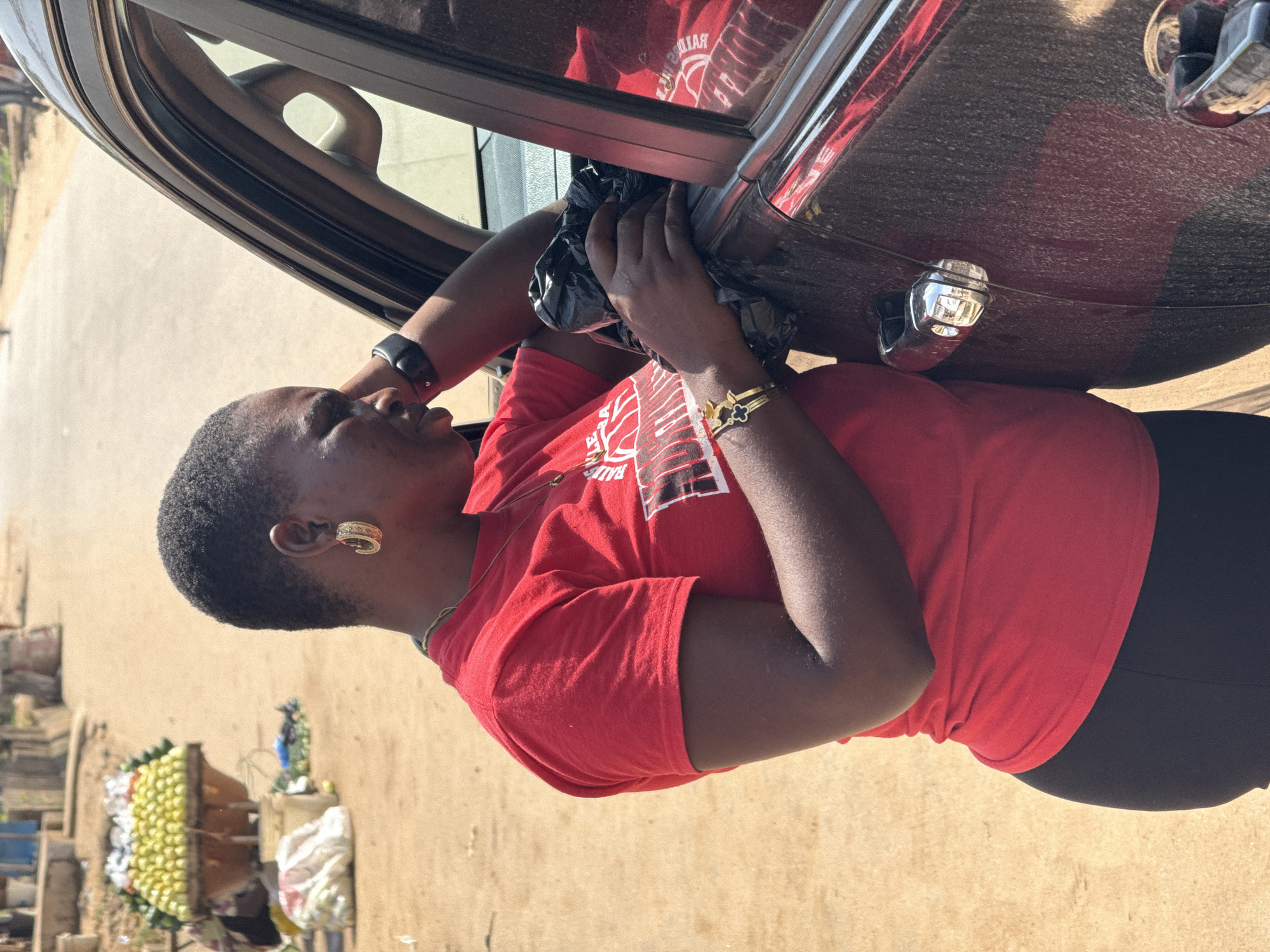 A medium shot captures a Black woman with short hair and gold hoop earrings standing next to the open passenger door of a dark-colored SUV. She wears a red t-shirt with white text and black leggings. Her right arm rests on the car, and she holds a crumpled black plastic bag in her hands, which are positioned near the car door. She appears to be looking down and to her right.
The background suggests an outdoor setting, possibly a street market. A cart filled with produce, including what appear to be yellow fruits, is visible in the left portion of the frame. Several white bags are also on the ground. The ground is a dusty, sandy color, and a paved road stretches into the distance, with hints of buildings and people in the background, indicative of a busy urban environment. The overall scene is consistent with a street market in Abuja, Nigeria, where vendors often sell goods from carts and the ground is unpaved in many areas.