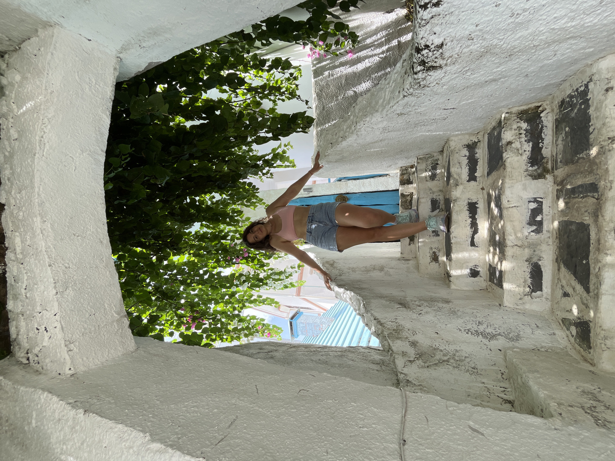 A young woman with dark hair, wearing a pink crop top and denim shorts, is lying on a narrow, white-washed stone staircase. She is looking towards the camera with a slight smile. Lush green foliage with some pink and white flowers hangs over the staircase from above. Sunlight filters through the leaves, casting dappled shadows on the rough, textured stone walls. The staircase appears to be located in a narrow alleyway or courtyard. In the background, hints of blue and white buildings can be seen, along with striped awnings. The overall impression is a sunny, warm day, with the woman enjoying a relaxed moment in a picturesque, possibly Mediterranean or Southeast Asian, setting.