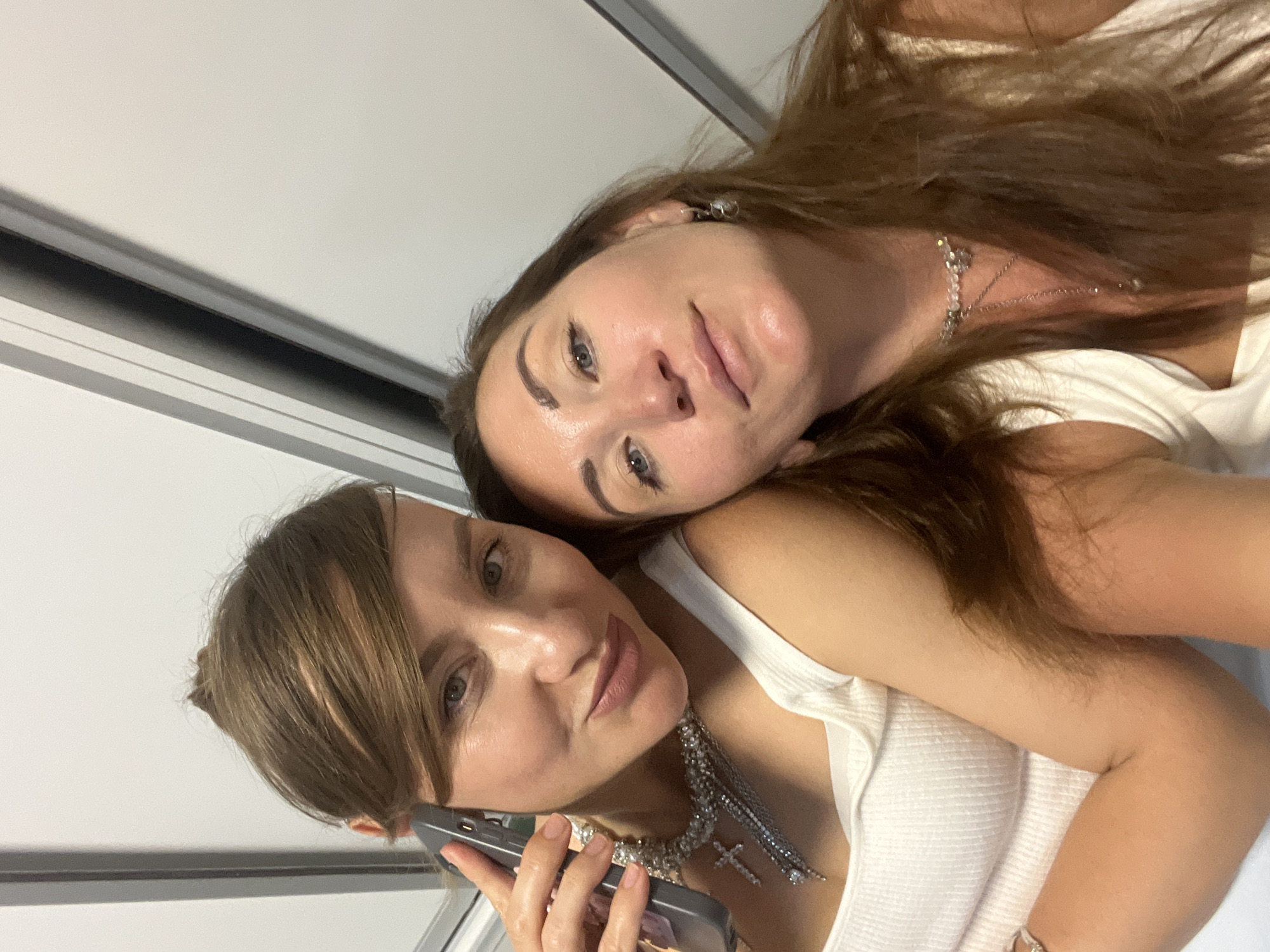 Two women are in a selfie taken indoors. The woman on the left is holding a phone to her ear and looking at the camera with a slight smile. She is wearing a white ribbed tank top and a sparkly necklace with a cross pendant. The woman on the right is leaning her head on the woman on the left's shoulder and is also looking at the camera. She has long brown hair and is wearing a white tank top and a delicate necklace. The background appears to be a wall with vertical elements, possibly a hallway or interior of a building. There is no indication of the city of Sochi, Russia in the image.