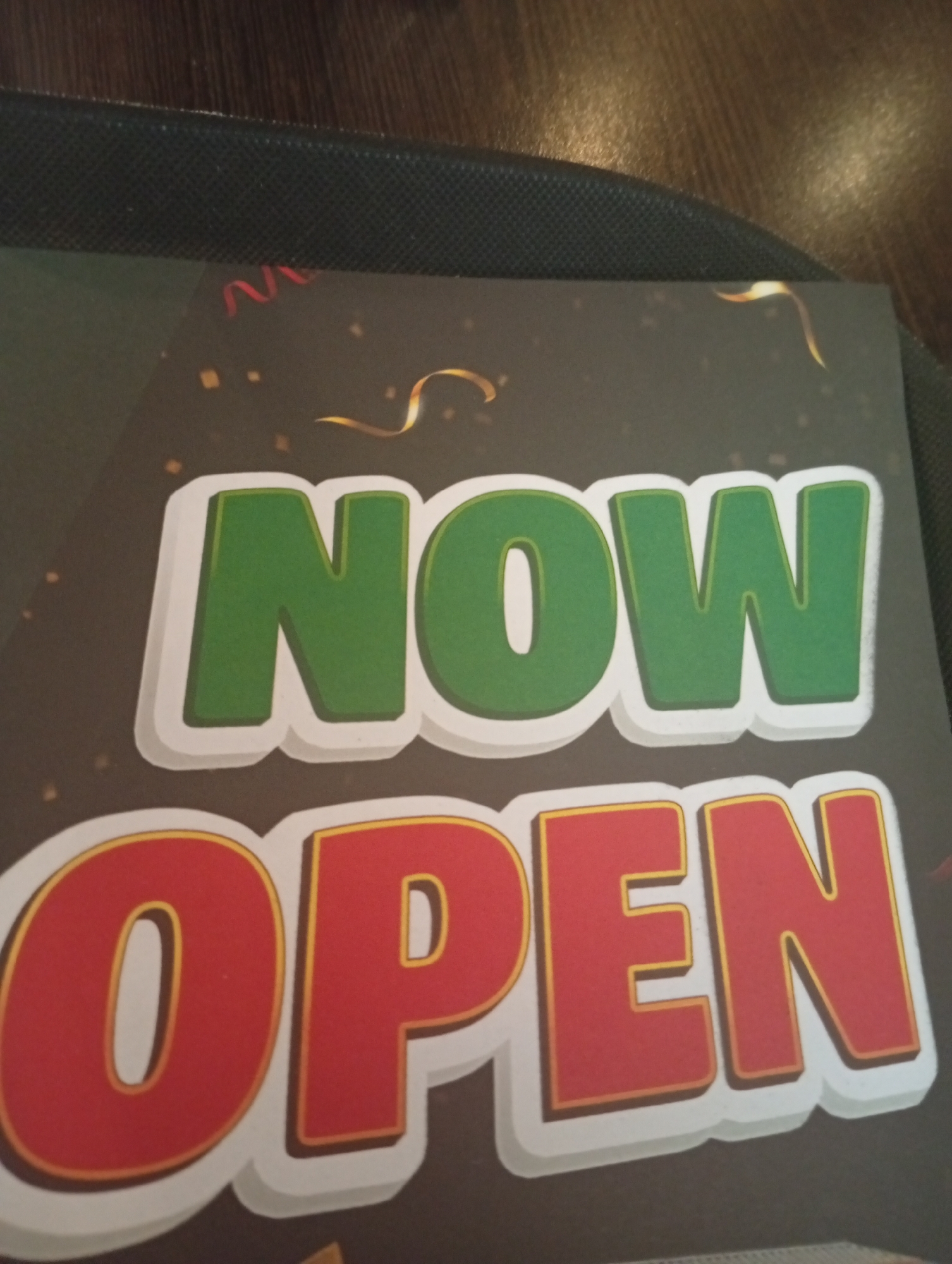 The image shows a close-up of a sign with the words "NOW OPEN" in large, bold letters. The word "NOW" is in green with a white outline, and the word "OPEN" is in red with a yellow outline and a white shadow. The sign has a dark background with scattered gold and red confetti. The sign appears to be on a table, with a dark, textured surface visible in the background. There is no visible location information in the image itself that would indicate it is in Al Rayyan, Qatar.