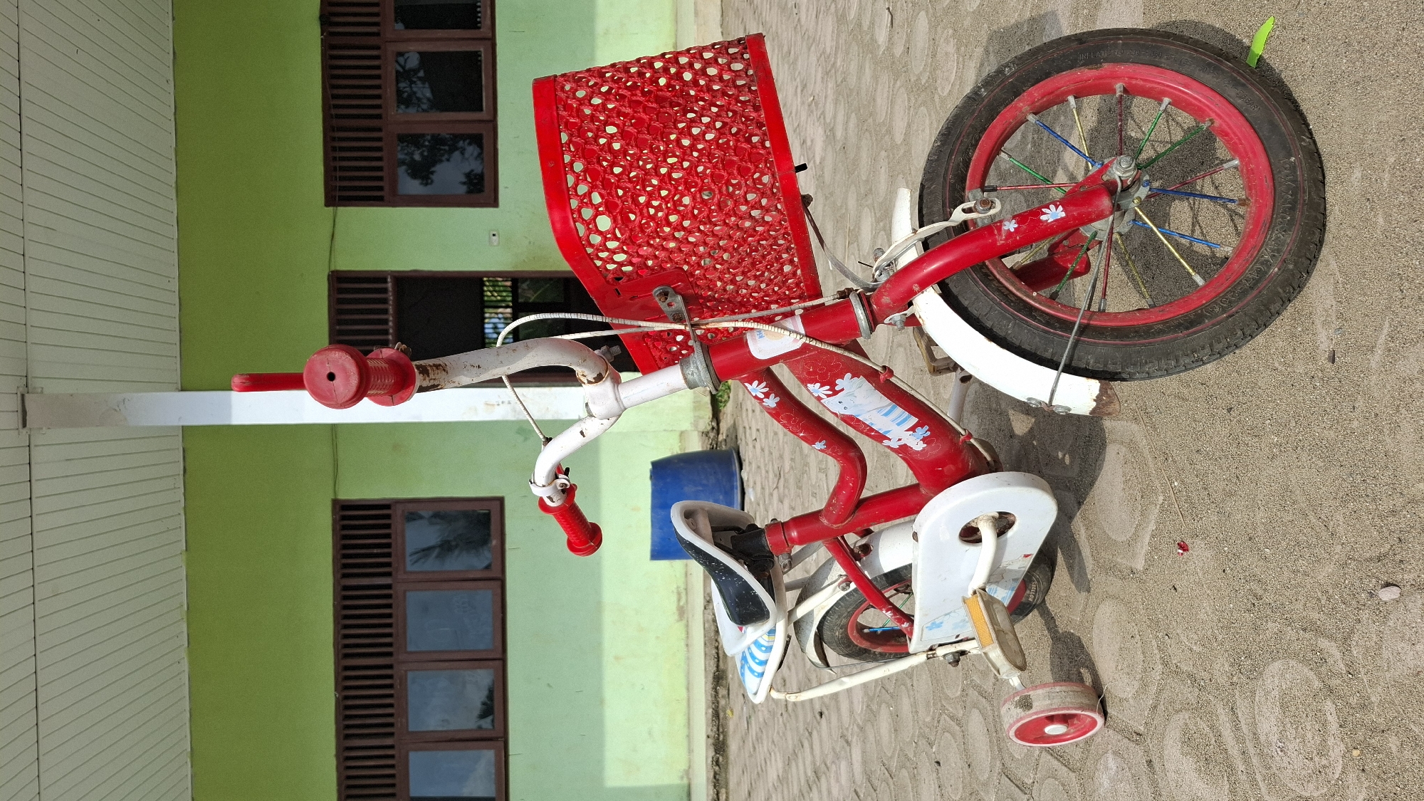 A bright red children's bicycle with white accents and a red basket is parked on a sandy ground in front of a light green building with dark brown windows. The bicycle has training wheels on the left side. The building has a white overhang and a white pillar supporting it. A blue barrel is visible behind the bicycle. The scene appears to be outdoors, possibly in a schoolyard or community area. The location is identified as Grong-grong, Indonesia.
