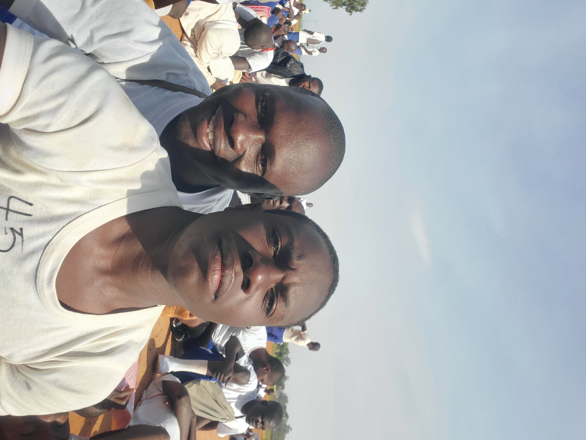 In Barakin Rafin Gora, Nigeria, two young men of African descent are the primary subjects of this self-taken photograph, positioned in the foreground. They are both wearing white t-shirts. The man on the left is smiling broadly, his mouth open, revealing teeth. The man on the right has a neutral expression. Both men appear to be outdoors during the daytime, with a clear sky in the background. The environment suggests a gathering or event, as other individuals, also appearing to be young men, are visible in the mid-ground and background. Many of these individuals are also wearing white t-shirts and are seated or standing on the ground. The ground appears to be sandy or dusty. The overall scene conveys a sense of community or group activity in an open, outdoor setting.