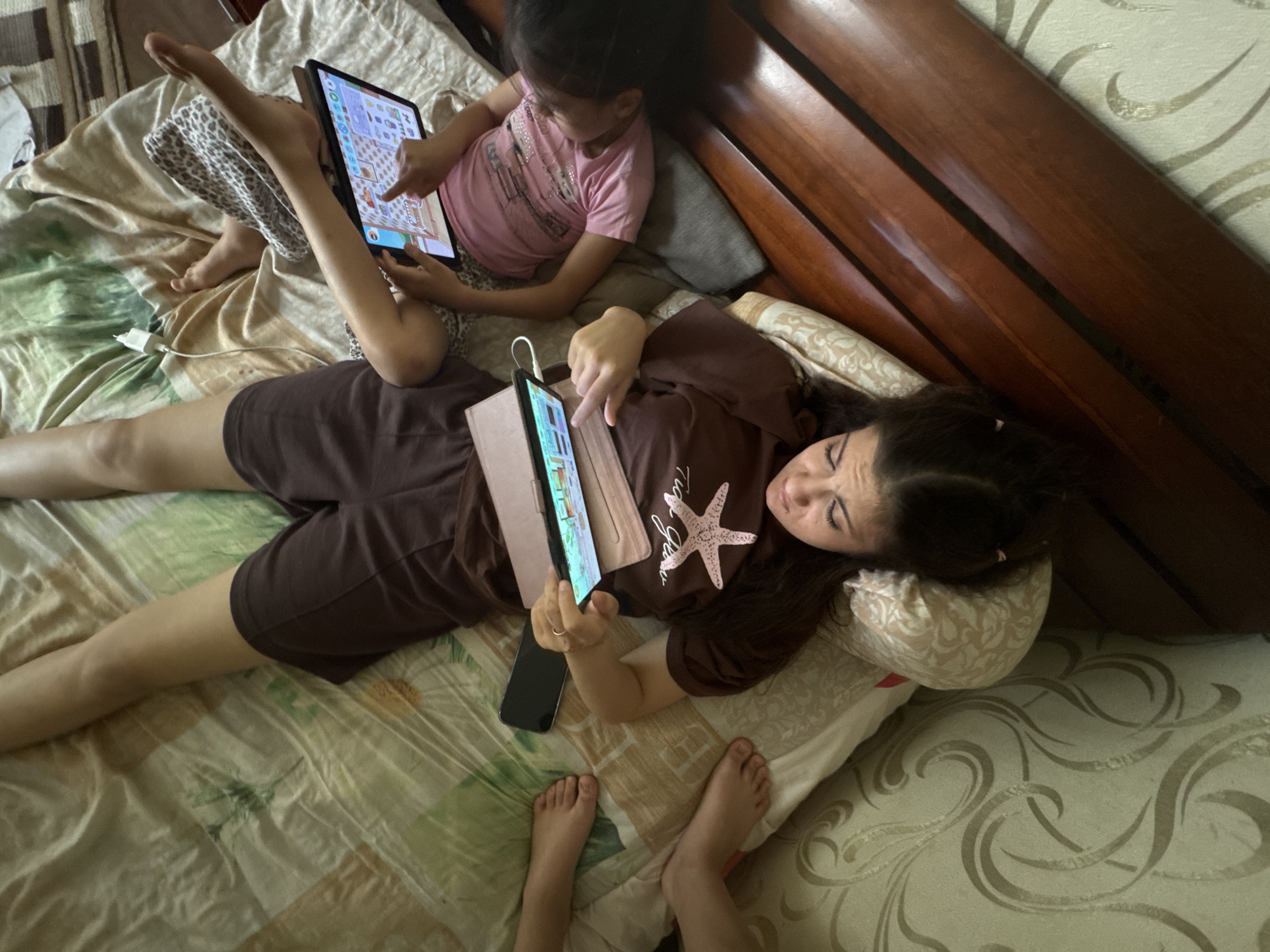 In an indoor setting, likely a bedroom, three individuals are positioned on a bed in the city of Unknown, Unknown. An adult female, with long dark hair, lies on her back with her head on a patterned pillow. She wears a dark brown t-shirt featuring a light pink starfish graphic and dark shorts. She holds a tablet, encased in a pinkish-brown folio cover, with both hands, viewing its screen which displays a colorful graphical interface. A gold-colored ring is visible on her left ring finger. A black smartphone rests on the bed next to her.
To her right, a juvenile individual, presumably female, lies on her back, wearing a pink t-shirt. She holds a larger tablet with both hands, also viewing its screen, which shows a brightly colored game or application interface. Her lower legs are partially covered by a white and black leopard-print blanket or garment. A white power adapter and cable are visible on the bed near her feet.
To the left of the adult female, only the lower extremities (feet and calves) of a third, juvenile individual are visible on the bed.
The bed is covered with a patterned duvet or comforter featuring sections of green, brown, and light-colored fabric. A wooden bed frame or headboard is partially visible along the right edge of the scene. All visible individuals are engaged with electronic devices.