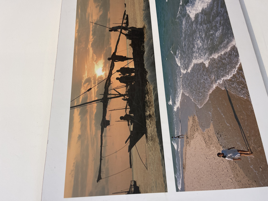 The image displays two distinct photographic prints, horizontally oriented, mounted on a white, possibly textured, wall or board. The overall composition is captured from a slightly elevated and angled perspective, revealing the white surface above and to the sides of the prints.
**Upper Print (Bububu, Tanzania - Coastal Scene at Sunset/Sunrise):**
This print depicts a coastal scene featuring a traditional wooden sailing vessel, likely a dhow, in the waters off Bububu, Tanzania, at either sunset or sunrise. The sky is dominated by warm orange and yellow hues, with scattered dark clouds silhouetted against the bright low-hanging sun on the right side of the frame. The water is relatively calm, reflecting the sky's colors. The main vessel, dark in silhouette, occupies the central midground. It features a prominent mast with rigging and a long boom extending horizontally. Approximately 6-8 individuals, also rendered in dark silhouette, are visible on and around this boat. One person stands near the bow, another on a spar extending from the mast, and others are grouped within the boat's main body. A visible wake or small waves are forming around the boat's hull. In the background, to the left and right, several other smaller, silhouetted boats are present on the water.
**Lower Print (Bububu, Tanzania - Beach Scene):**
This print shows a wide, sandy beach in Bububu, Tanzania, during daylight hours. The ocean occupies the right side of the frame, with turquoise water and multiple lines of white-capped waves breaking and receding on the shore. The beach sand is light brown, with darker, wet areas near the water's edge. Two individuals are present on the beach. In the lower-left foreground, a female figure, wearing a white t-shirt and light-colored shorts, walks barefoot towards the camera. Her shadow extends behind her on the sand. Further up the beach, closer to the water and towards the center-right, a male figure, dressed in dark clothing (possibly shorts and a t-shirt), walks barefoot away from the camera. The general environment is open and unpopulated beyond these two individuals.