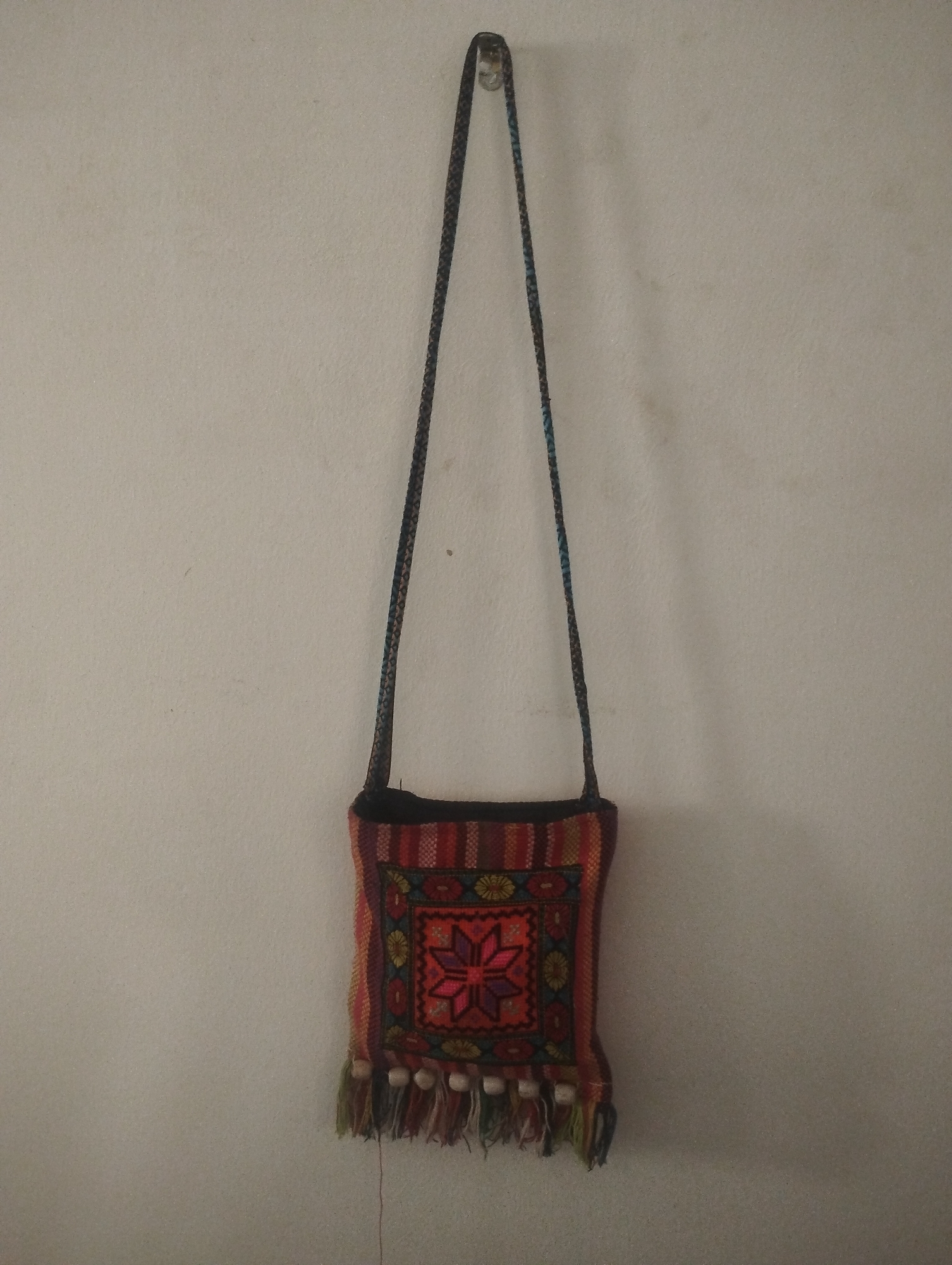 A square, hand-stitched shoulder bag hangs from a metal hook against a plain, off-white wall. The bag is primarily shades of red, with a central geometric floral design, and a fringed bottom edge with small white beads interspersed. The strap is long and braided. The location of the image is Dutsen Alhaji, Nigeria.