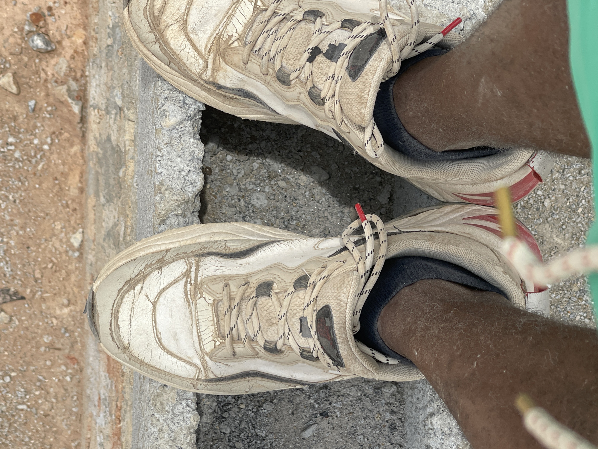 A high-angle, close-up shot shows a person wearing dirty, white sneakers standing on a concrete surface. The legs of the person are visible, showing dark skin tone and wearing dark socks. The shoes appear worn and aged, with cracks and dirt marks. The background shows a concrete ledge and a patch of dirt or unpaved ground. Based on the prompt, the location is Angwan Sayawa, Nigeria, but this cannot be confirmed from the image. The person appears to be stationary, standing on the concrete. The image is focused on the shoes and legs of the person, with a view of the ground surface.