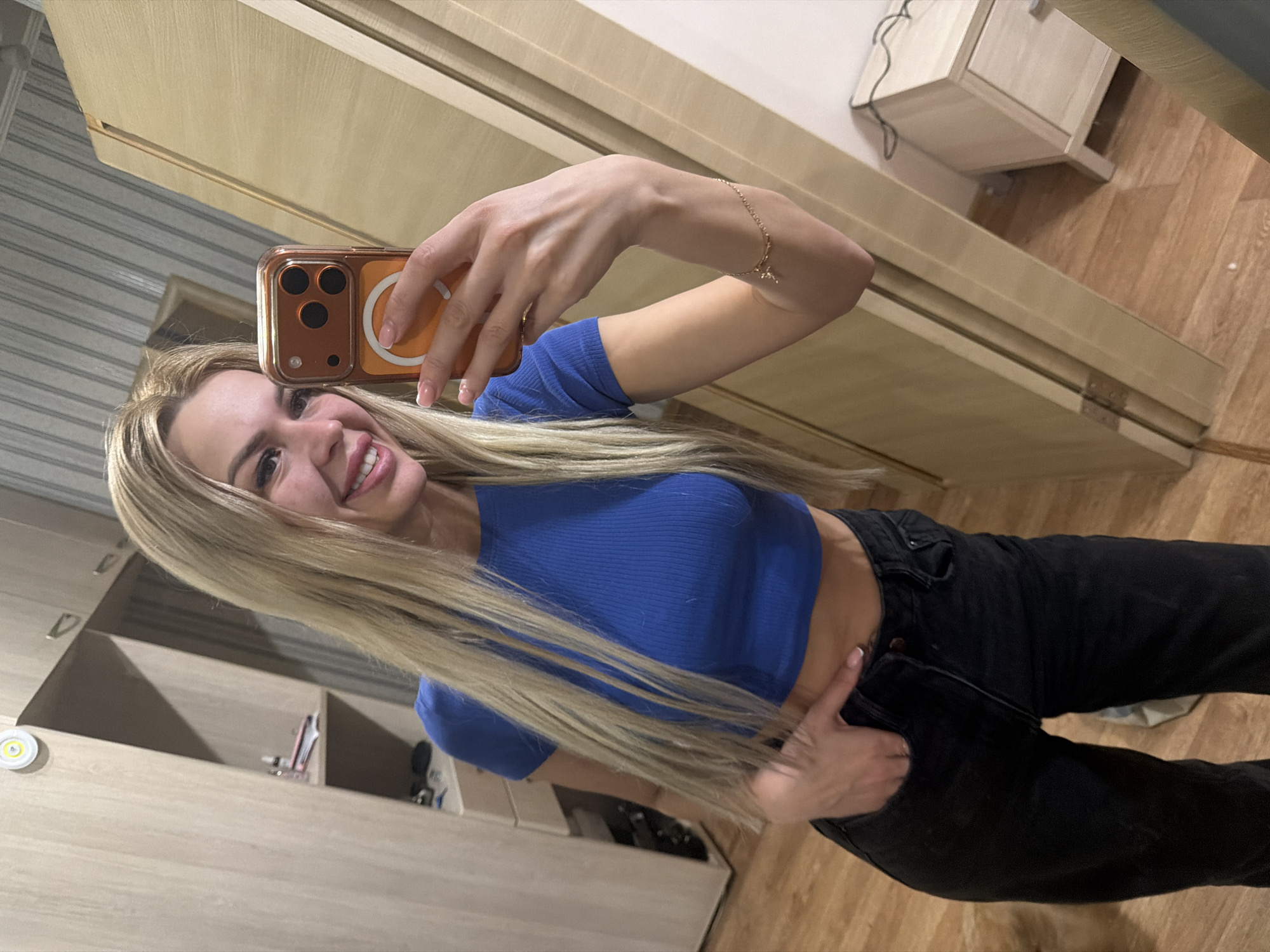 A young woman with long blonde hair, wearing a blue ribbed crop top and black jeans, takes a selfie in a room with light wood furniture and flooring. She is holding an orange-cased smartphone in her right hand, with her left hand in her pocket. A gold bracelet with a cross charm is visible on her left wrist. The background includes a closet, a drawer unit, and a door. The overall scene appears to be a residential interior.