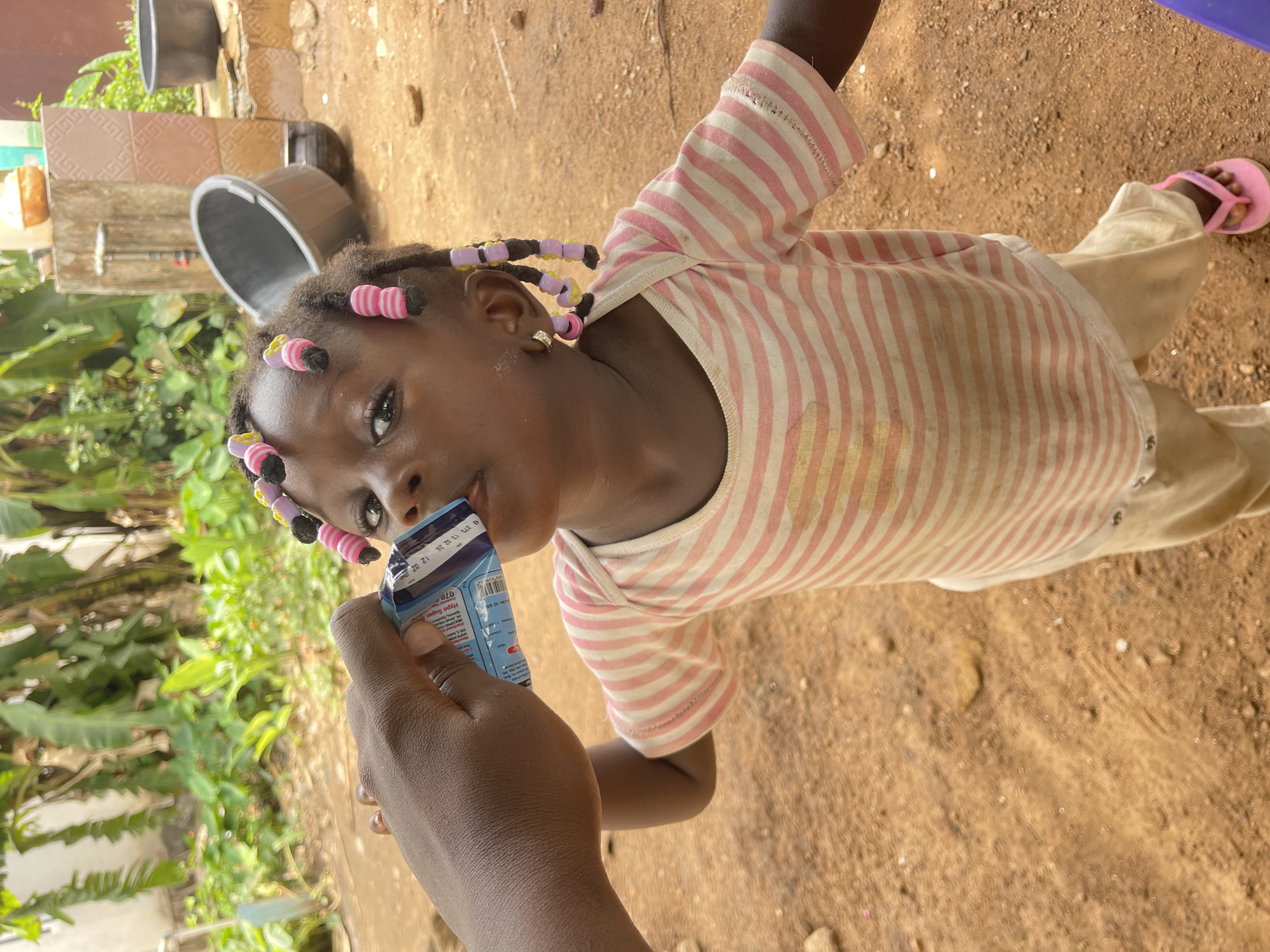 The image, oriented sideways, presents an outdoor scene featuring a young child (likely a girl) actively drinking from a juice box, which is held by an adult's hand. The child, with dark skin, is centered in the frame, looking directly towards the camera with a focused expression.
She has black hair styled in numerous braids, each adorned with colorful plastic beads in pink, yellow, and purple. Her attire consists of a light-colored, short-sleeved top featuring thin pink vertical stripes, paired with light-colored trousers. A small golden hoop earring can be seen in her right ear, and she wears pink flip-flops on her feet.
An adult's dark-skinned hand is prominently shown holding a blue and white juice box, with the straw positioned in the child's mouth. Visible text on the juice box includes the brand "Hypo Super" and numerical details such as "070", and date-like sequences like "27/11 12:02" and "25 12/02", possibly indicating production or expiry information.
The setting is an outdoor, daytime environment. The ground is a reddish-brown dirt, suggesting a natural, unpaved area. Lush green vegetation, including large leaves, is visible on the left side of the background. Further back, dark-colored buckets or basins and a tiled surface with a geometric pattern are discernible, hinting at a functional outdoor area. The bright, natural lighting indicates clear weather. The scene portrays a simple, everyday moment, consistent with the specified location of Akwanga, Nigeria.