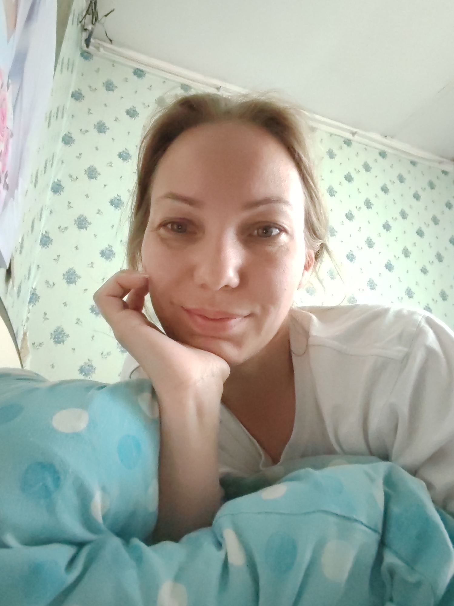 The image shows a young woman with blonde hair and brown eyes, lying in bed and resting her chin on her hand. She is wearing a white t-shirt and is partially covered by a blue polka-dotted blanket. The background consists of light green wallpaper with a floral pattern and a white ceiling. The lighting appears to be natural, coming from an unseen source. The woman is looking directly at the camera with a soft smile. The overall impression is one of a casual, at-home moment. There is no information available to indicate the location in the city of Чухлэм, Russia.