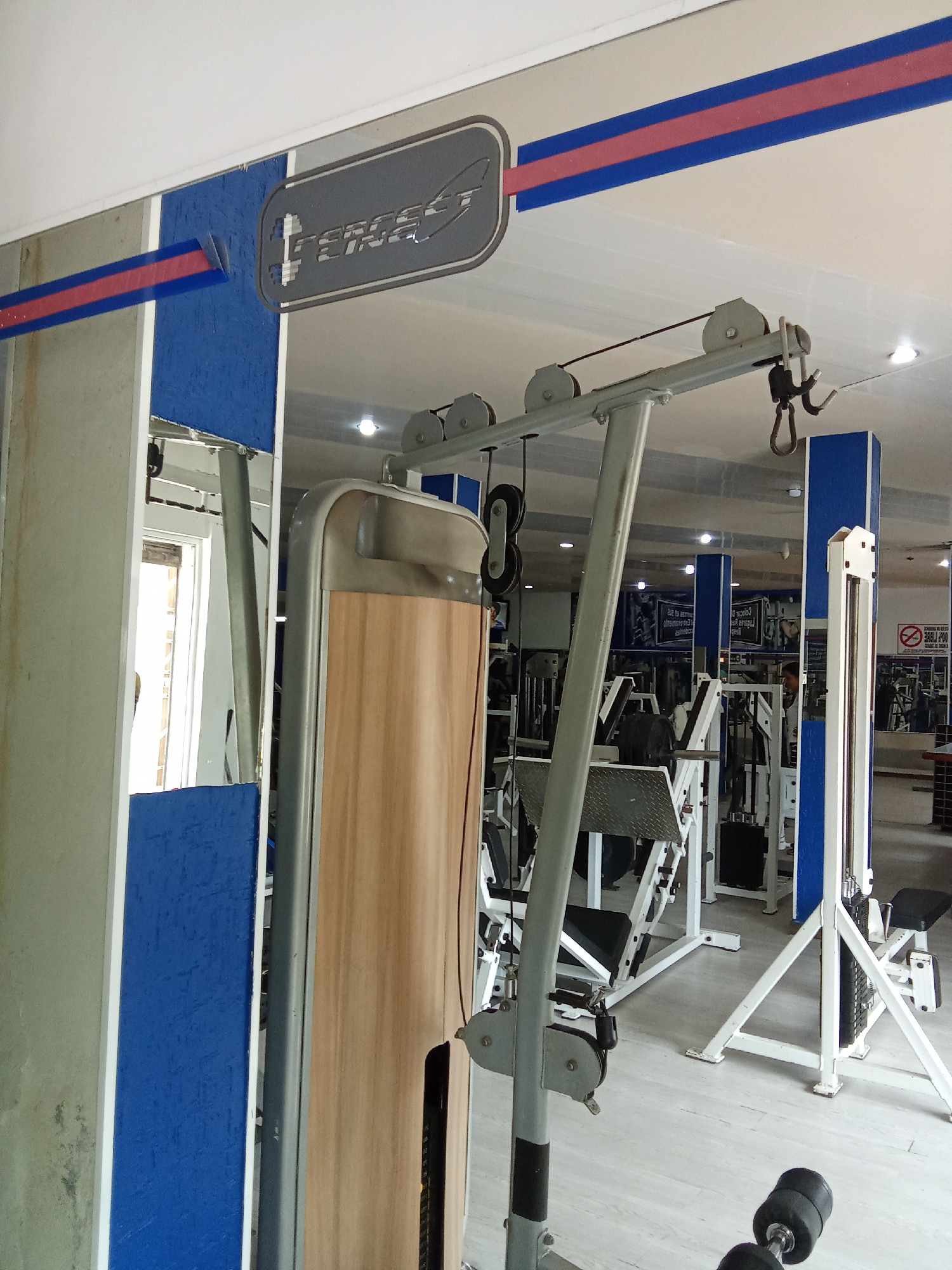 The image depicts the interior of a gym in Altagracia, Venezuela. A gray and light brown weight machine is prominent in the foreground. In the background, various other gym equipment pieces, including a white and black leg press and a lat pulldown machine, are visible. The walls and columns are painted with blue and white accents, with a red stripe near the ceiling. A dumbbell lies on the light gray floor near the camera. The lighting suggests a bright interior space.