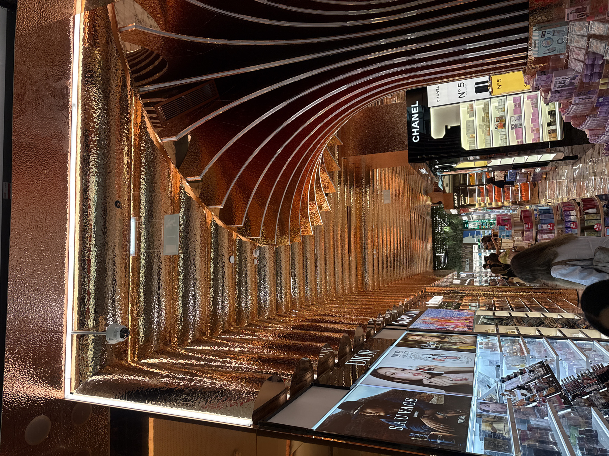 The image displays an opulent indoor retail environment, likely a high-end perfume and cosmetics section within a store in Odivelas, Portugal. The dominant feature is a striking architectural design composed of a curved, tunnel-like structure crafted from highly textured, reflective copper or bronze-toned material. This surface, resembling hammered metal, gleams under bright strip lighting integrated along its length, creating a luxurious and dynamic visual effect.
Along the right side of the scene, numerous well-lit display units showcase a wide array of perfumes and cosmetic products. A prominent black display unit featuring the brand "CHANEL" and its iconic "N°5" perfume is clearly visible. Below the curved copper structure, large, illuminated digital displays feature advertisements for other luxury brands. Visible text includes "DIOR," "DIOR ADDICT," "SAUVAGE ELIXIR," and "GUERLAIN," indicating a selection of premium fragrances and beauty products.
Several shoppers are present, seen mostly from behind or partially, engaged in browsing the extensive product offerings. Their activity suggests a typical shopping experience in a beauty retail setting. The overall lighting is bright and artificial, enhancing the presentation of the products and the reflective qualities of the architectural elements. A security camera is also visible, mounted on a pole near one of the light strips. The atmosphere conveys modern elegance and a high-end shopping experience.