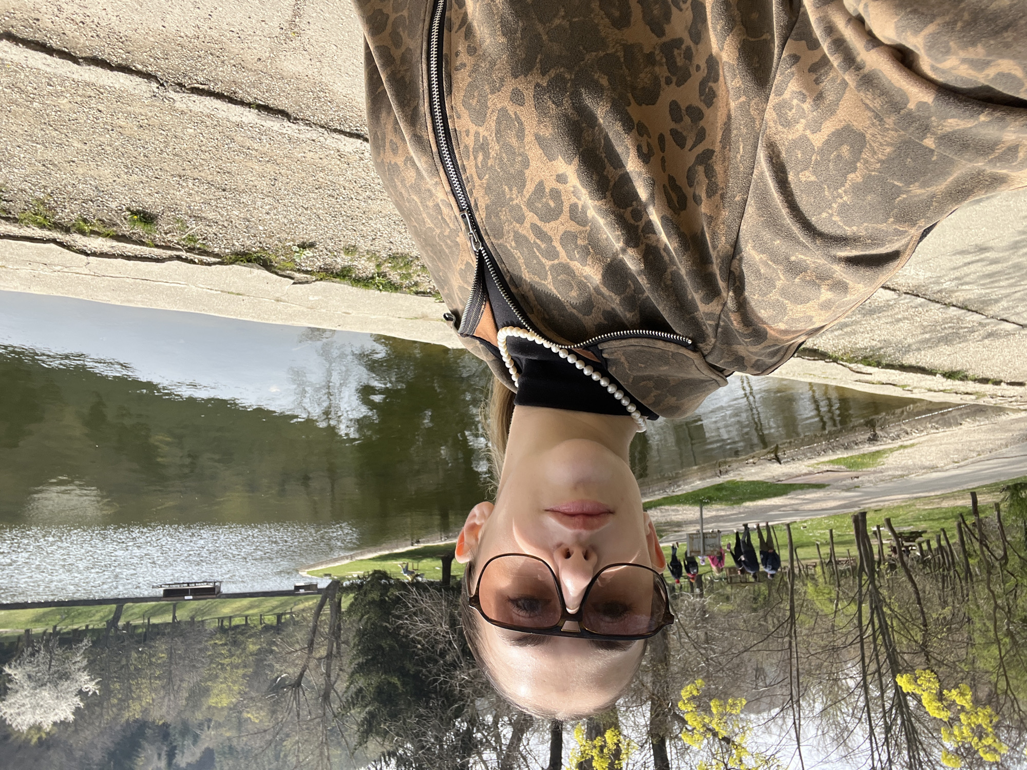 A person with glasses is facing the camera, wearing a brown animal print jacket with a silver zipper and a pearl necklace. They are standing beside a body of water, possibly a lake or pond. In the background, there are trees, green spaces, and what appears to be a group of people walking. There are also benches and other structures visible along the water's edge. This image was taken at Park Bachinovo, in Blagoevgrad, Bulgaria.
