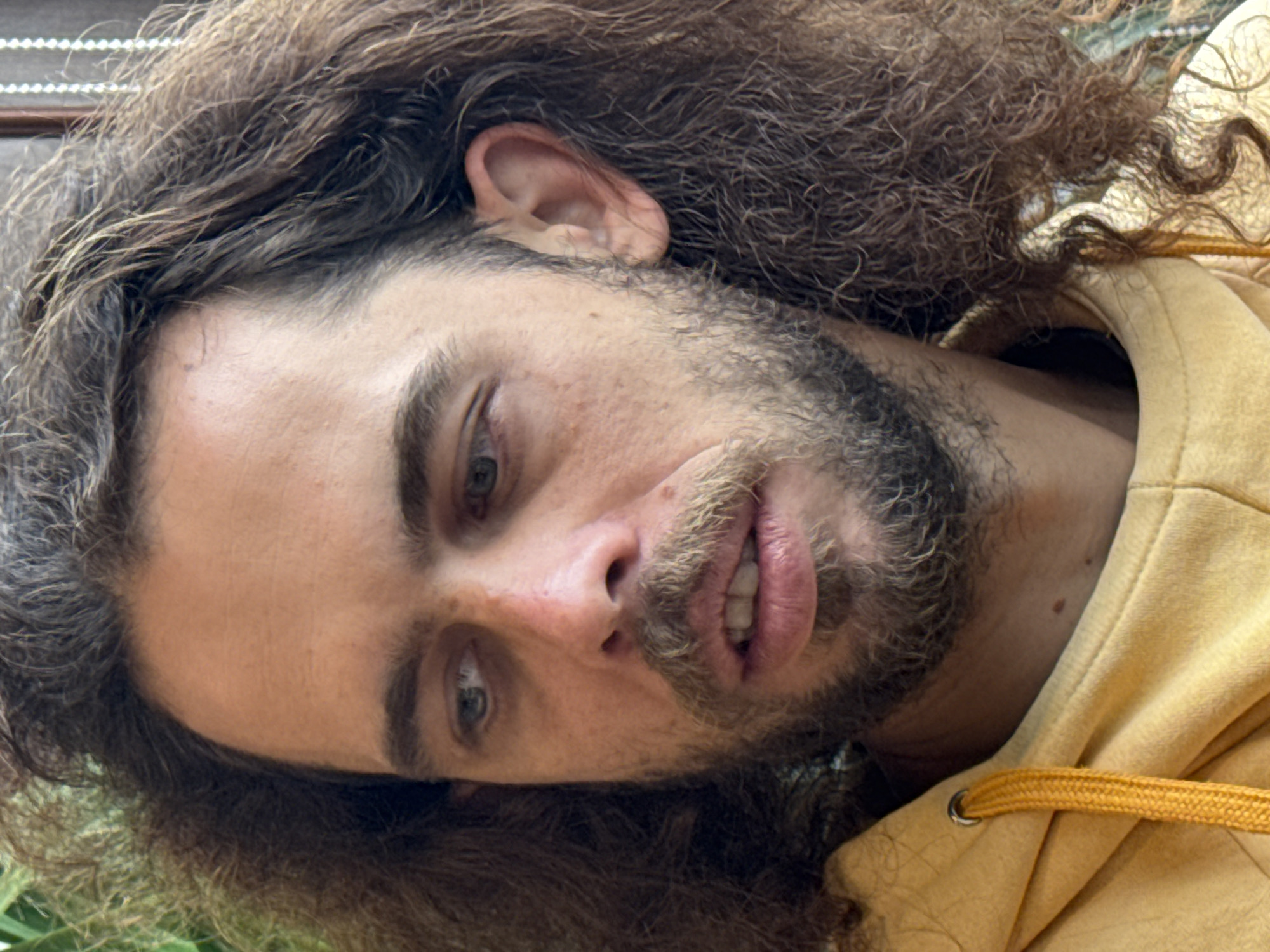 A close-up portrait of a man with long, curly brown hair and a light beard. The man is wearing a yellow hoodie and is looking slightly downward and to the left. The background is out of focus, with some greenery visible. The location cannot be determined as Binh Lu, Vietnam from the image alone.