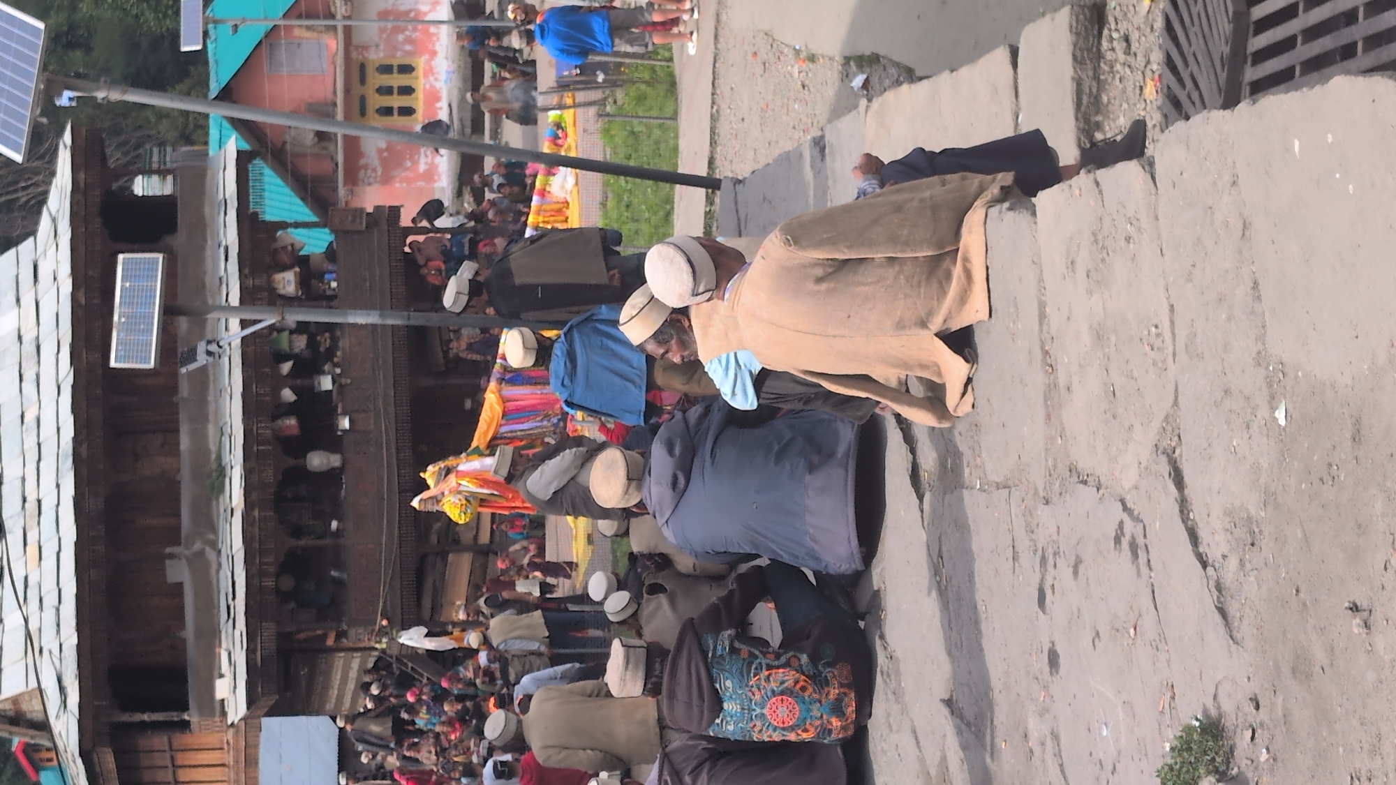 In Malana, India, a group of men are gathered outdoors, seated on a stone pathway in front of traditional wooden buildings. The architecture features multi-tiered wooden structures with ornate carvings and balconies. The men, dressed in traditional attire including caps, are observing an event or ceremony. In the background, colorful fabrics are displayed, suggesting a festive or cultural occasion. Solar panels are visible on poles, indicating a blend of traditional and modern infrastructure. The environment appears to be a village setting with cobblestone paths and surrounding buildings.