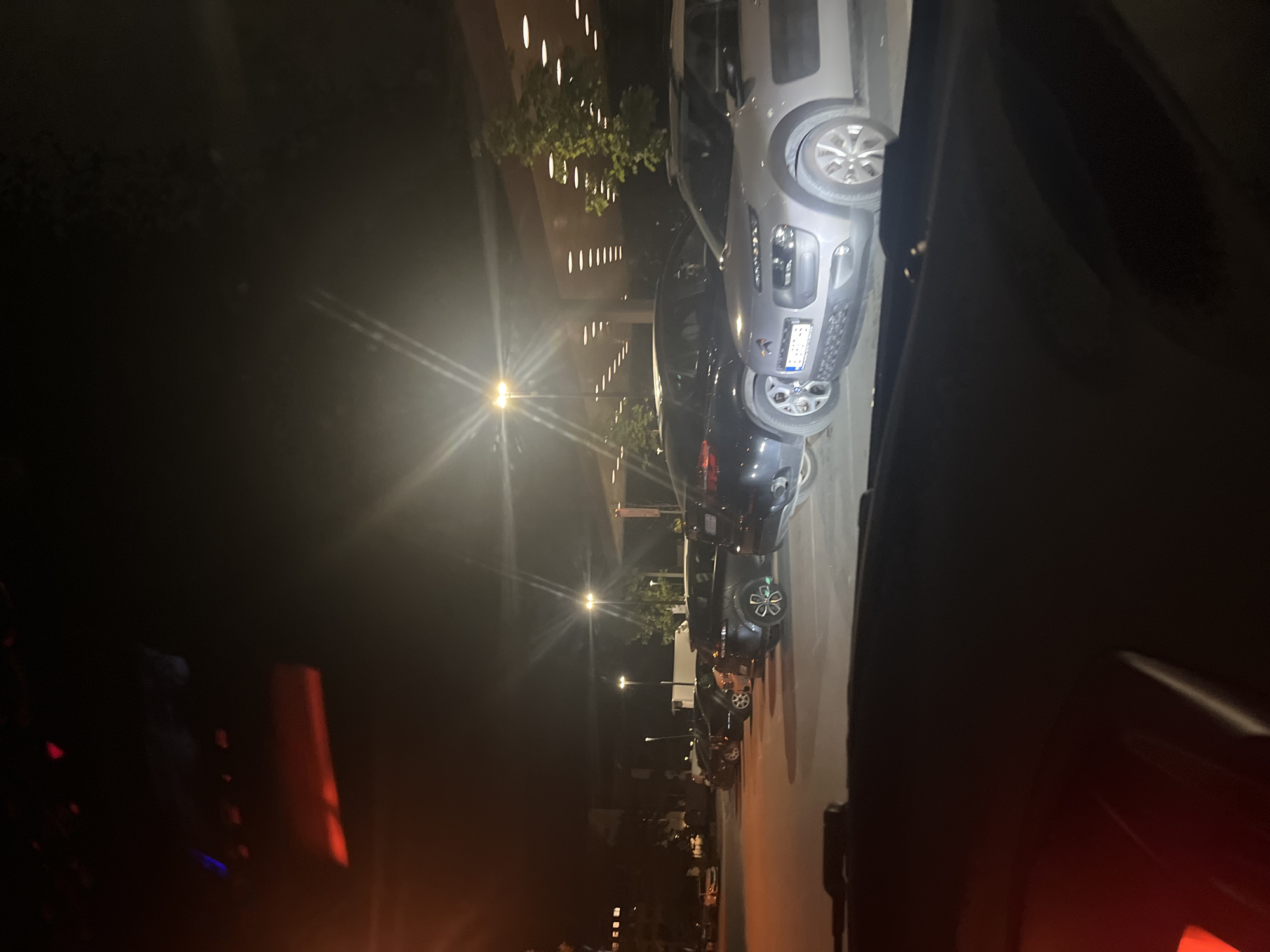 The image, taken at night from inside a vehicle, depicts a street or parking area in an urban setting. The perspective is from a car, with elements of the interior, such as parts of the dashboard and a door frame, visible on the left and right edges.
On the right side of the frame, several cars are parked parallel to the road. The most prominent vehicle is a light grey SUV, possibly a Fiat 500X, with its front facing towards the right. Its license plate, which appears to be Bulgarian (starting with "PB"), is visible but partially obscured by glare. Another dark-colored car is parked behind it, and more vehicles are visible further down the street.
On the left side, a multi-story building stands, characterized by numerous uniformly spaced, vertically elongated illuminated features that suggest either windows or integrated architectural lighting. Some foliage or trees are visible in front of the building.
The scene is predominantly dark, illuminated by several bright streetlights positioned along the street, which cause significant lens flares. These flares appear as radiant starbursts and streaks across the image. The artificial lighting creates strong contrasts, with much of the background receding into deep shadow. The overall impression is that of a quiet, illuminated street at night. No people or active events are discernible.