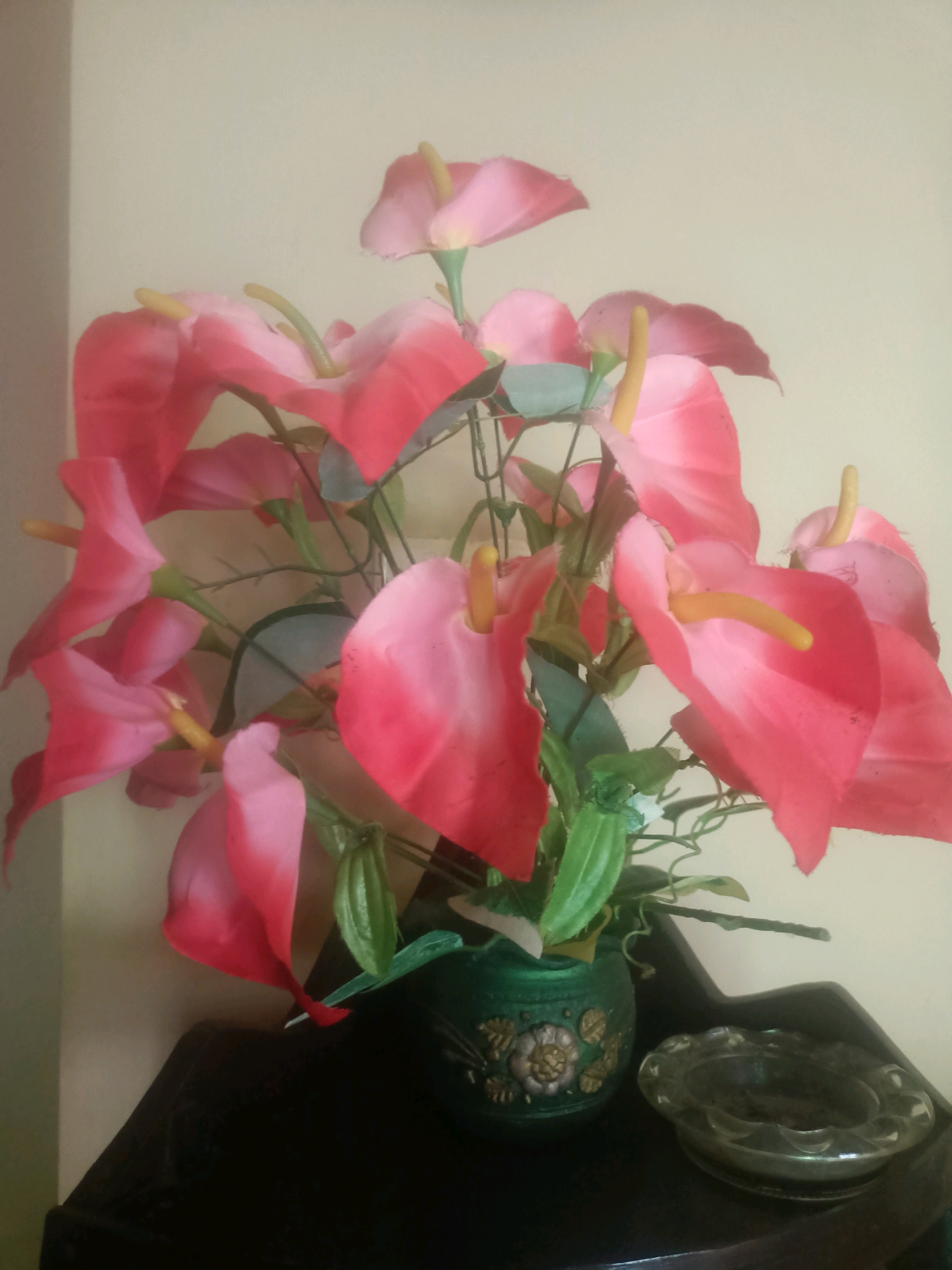 A bouquet of artificial pink anthurium flowers is arranged in a green vase with a floral pattern. The vase sits on a dark wooden surface, to the right of which is a clear glass ashtray. The background is a plain, light-colored wall. The image was taken indoors. The location is not specified in the provided information.