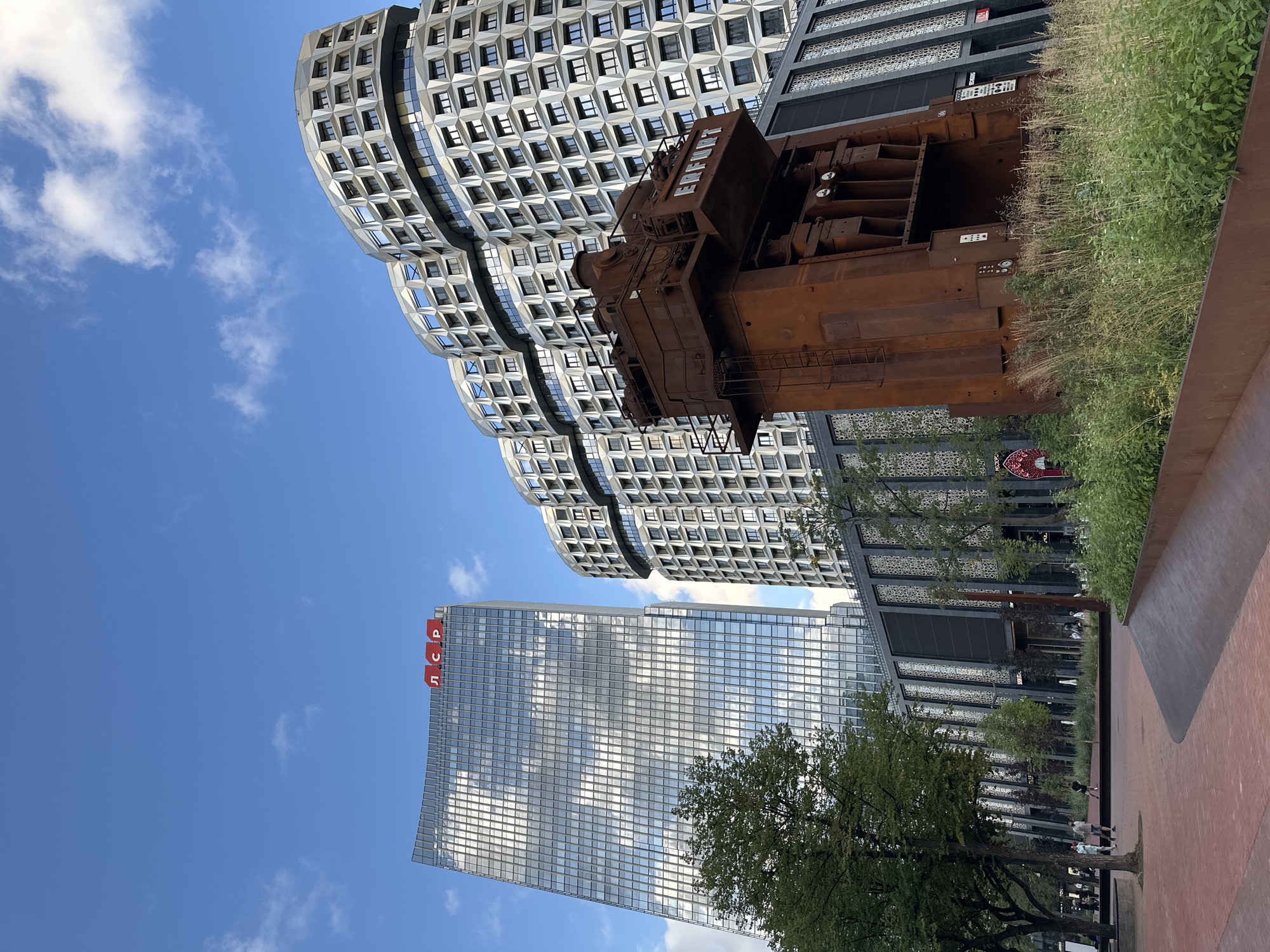 The image shows a bright, sunny day with a blue sky and scattered clouds. In the foreground, a reddish-brown paved area curves to the right. To the left, a tree with green leaves stands in front of a brick walkway. Beyond the walkway, a modern glass skyscraper with a red logo at the top, reading "ЛСР", dominates the left side of the frame. To the right of the skyscraper, two curved, modern buildings with intricate geometric facades rise against the sky. In the middle ground, a large, rusted industrial machine, possibly a press, is situated in front of a dark-paneled building. This structure has visible ladders and control panels. Green grass and plants grow around the base of the machine. In the background, more modern buildings and trees are visible, suggesting an urban environment. People are sparsely scattered throughout the scene, some walking on the pathways. The overall impression is of a contemporary urban landscape with an juxtaposition of old industrial art and modern architecture. The "ЛСР" logo on the skyscraper indicates the location is likely connected to the LSR Group, a Russian company, and based on architectural style and common landmarks, this is likely the LSR Building in Moscow, Russia, which features this prominent industrial art installation.