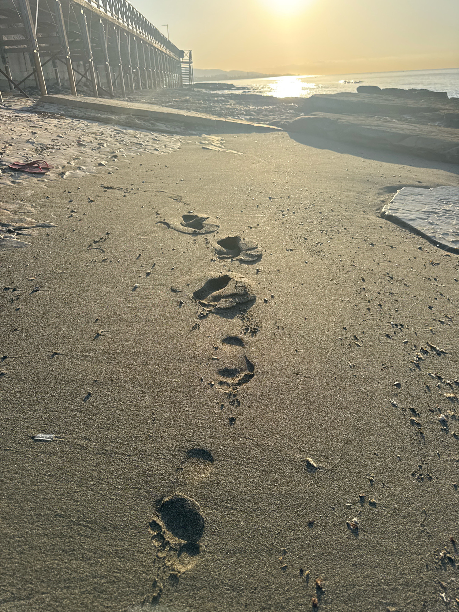 The image depicts a sandy beach at sunrise or sunset, with the sun casting a warm glow over the scene. Footprints are visible in the sand, indicating recent human presence. To the left, a wooden pier or boardwalk extends into the distance, with its support structure visible. The sea is calm, with gentle waves lapping at the shore. The environment appears to be a coastal area, possibly in Agios Tychonas Community, Cyprus, known for its beaches and seaside attractions. There are no people or animals visible in the frame.