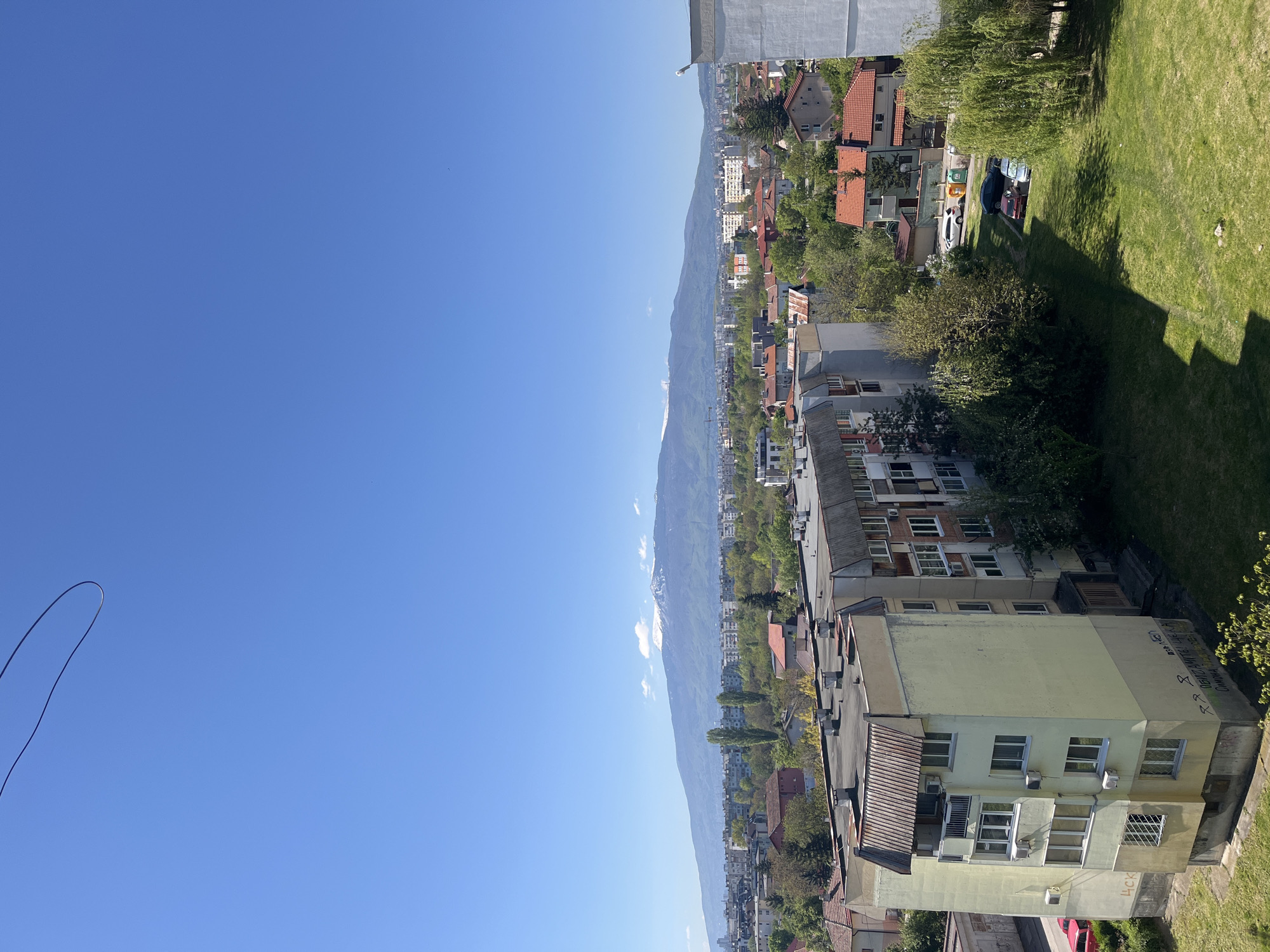 The image depicts a view of Sofia, Bulgaria, with a clear blue sky overhead. In the foreground are residential buildings with a mix of architectural styles, some featuring red tile roofs. Trees are interspersed among the buildings, adding greenery to the urban landscape. In the distance, a mountain range rises along the horizon, with some peaks visible through the haze. A patch of green lawn is visible in the upper right corner. A black wire or cable is visible against the blue sky in the upper left of the image.