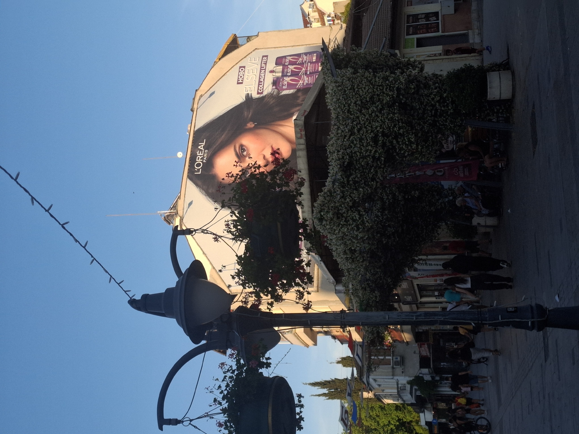 The image shows a street scene in a city under a clear blue sky. A large billboard featuring a woman's face and a L'Oréal Paris advertisement is prominently displayed on the side of a building. In the foreground, a lamppost with hanging flower baskets stands to the left, partially obscuring the view of the street. People are walking along the sidewalk, which is paved with stone tiles. Shops and buildings line the street, with trees and foliage adding to the urban landscape. The lighting suggests it is late afternoon or early evening, with the sun casting long shadows.