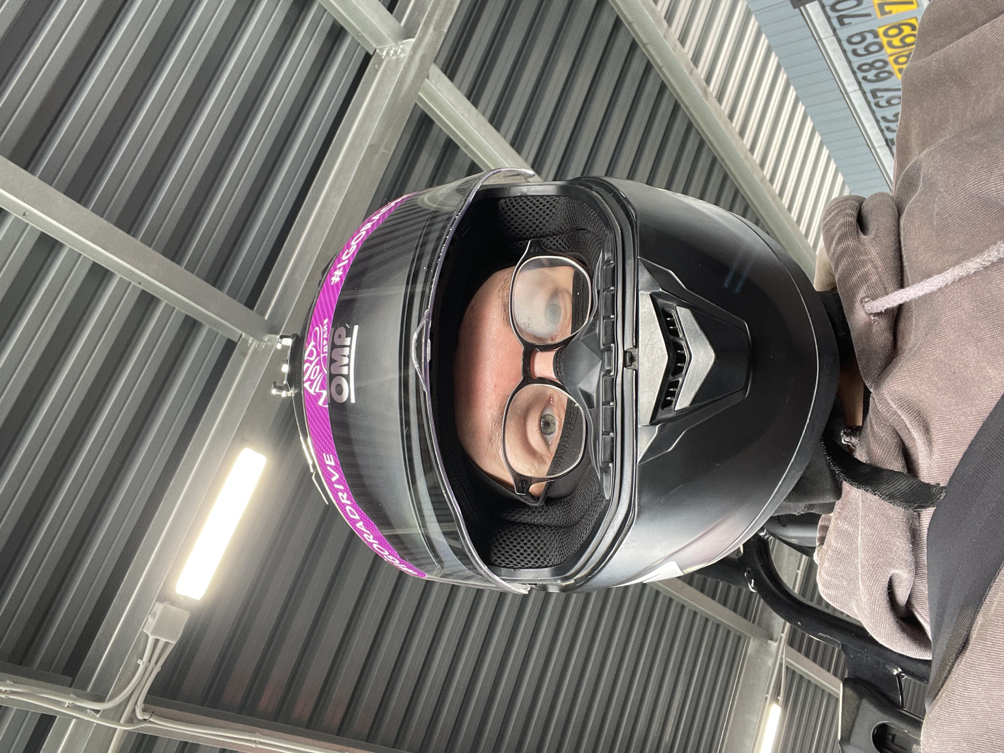 A low-angle, eye-level selfie shows a person wearing a black motorcycle helmet with a pink racing stripe and the OMP logo. The person is also wearing glasses and a black mask, with only their eyes visible. They are wearing a tan hoodie, and a black seatbelt is visible across their chest. The background consists of a corrugated metal roof and support beams, suggesting an indoor or covered outdoor structure, possibly a garage or a workshop. The image seems to have been taken at a racing or driving facility. The presence of numbers on a blue and yellow sign in the background, "67 68 69 70 69 7", is consistent with a racing pit lane or a parking area for vehicles. The location is identified as Novozhilovo, Russia.