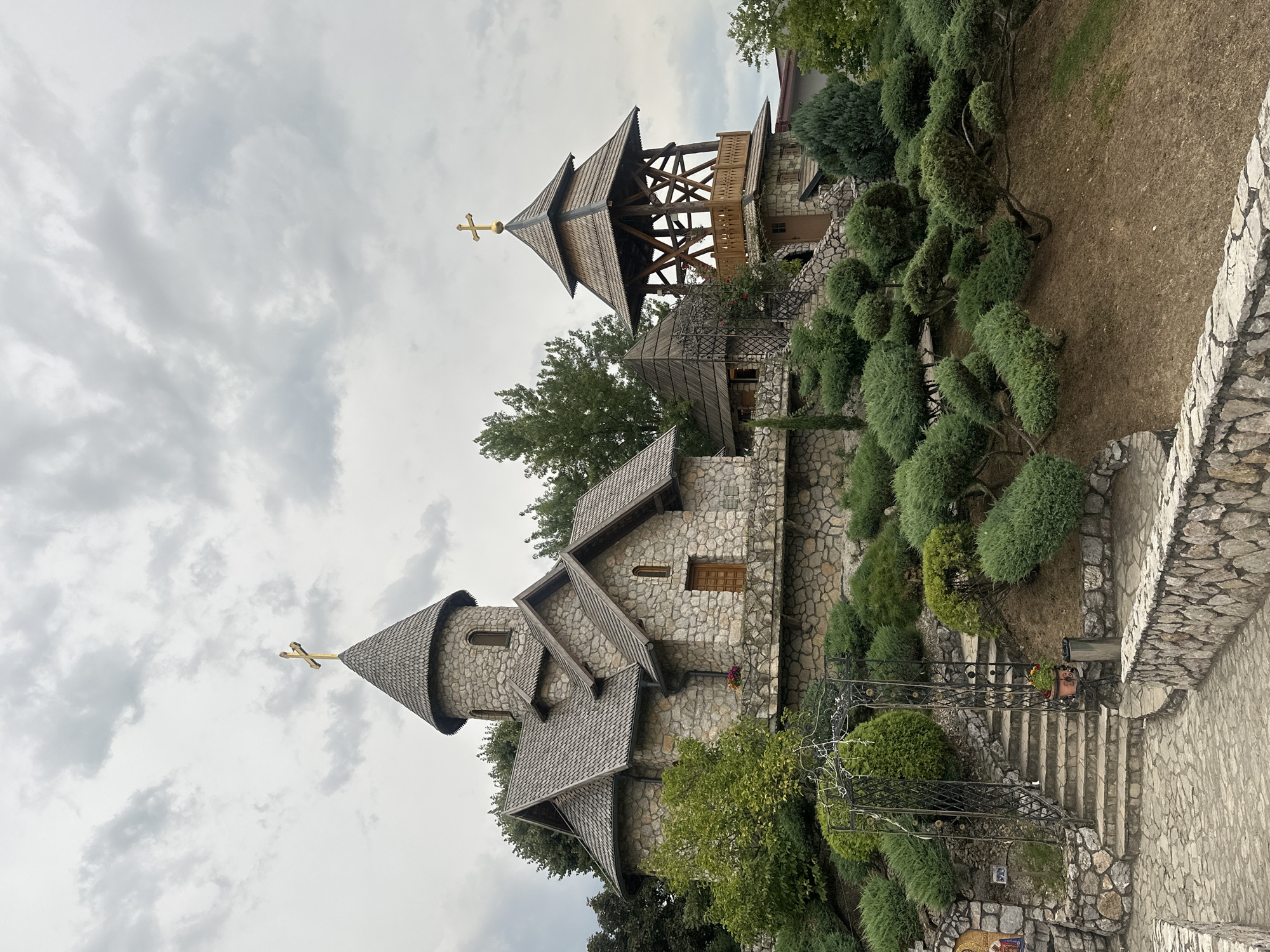 The image shows a stone church with a wooden bell tower, located on a hillside in the village of Dijelovi, Bosnia and Herzegovina. The church has a circular tower with a conical roof topped by a gold cross, and a rectangular section with a gabled roof. The bell tower is a multi-tiered wooden structure, also topped with a gold cross. The hillside is covered with manicured bushes and trees, and a stone pathway leads up to the church. The sky is overcast with grey clouds. There are no people visible in the image.