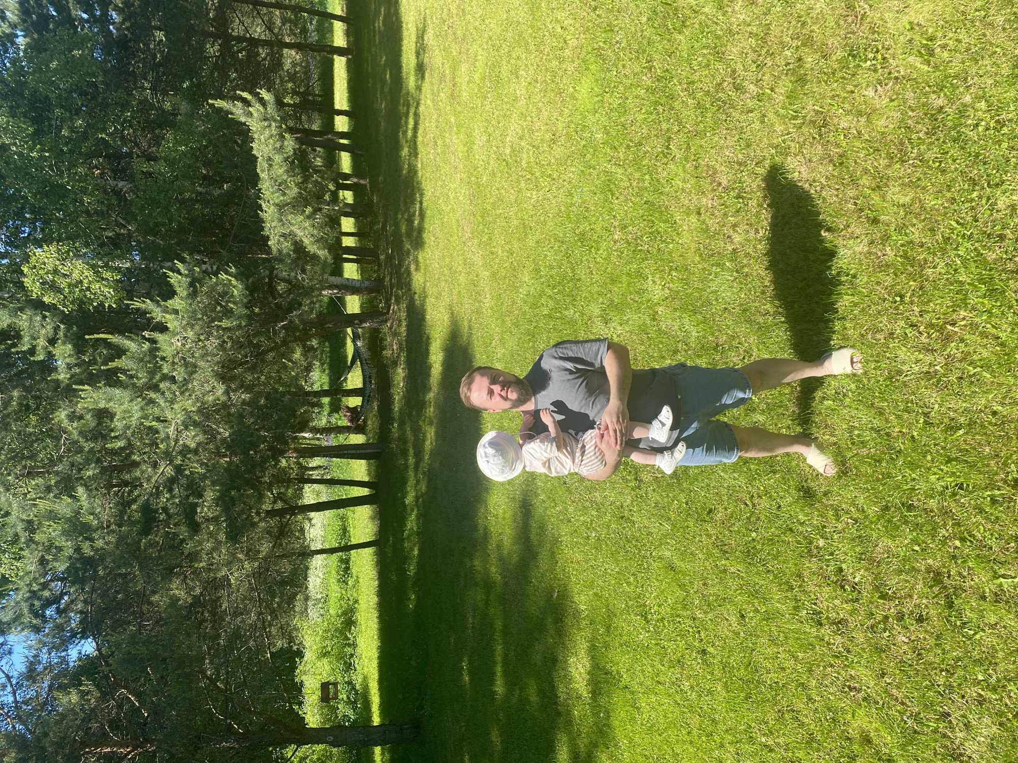 This image, captured in Копнино, Russia, displays an outdoor scene on a brightly lit, clear day, indicated by strong shadows suggesting midday or early afternoon. The foreground and midground are dominated by a lush, well-maintained green lawn or field. An adult male, appearing to be in his late 20s to mid-30s, stands centrally, facing the camera with a neutral expression. He has short dark hair and a short beard, wearing a dark grey short-sleeved t-shirt, blue denim shorts, and light-colored slip-on sandals. He holds a young child, estimated to be 1-2 years old, who is dressed in a white sun hat, a light-colored short-sleeved top, striped shorts, and light sandals. The child's back is partially towards the camera, and it faces towards the man's left. The background consists of a dense line of trees, primarily evergreens (pines) interspersed with some deciduous trees, casting shadows across the grass. A wooden bird feeder is visible hanging from a tree branch in the upper-left background, and a light-colored hammock is strung between two trees in the far-right background.