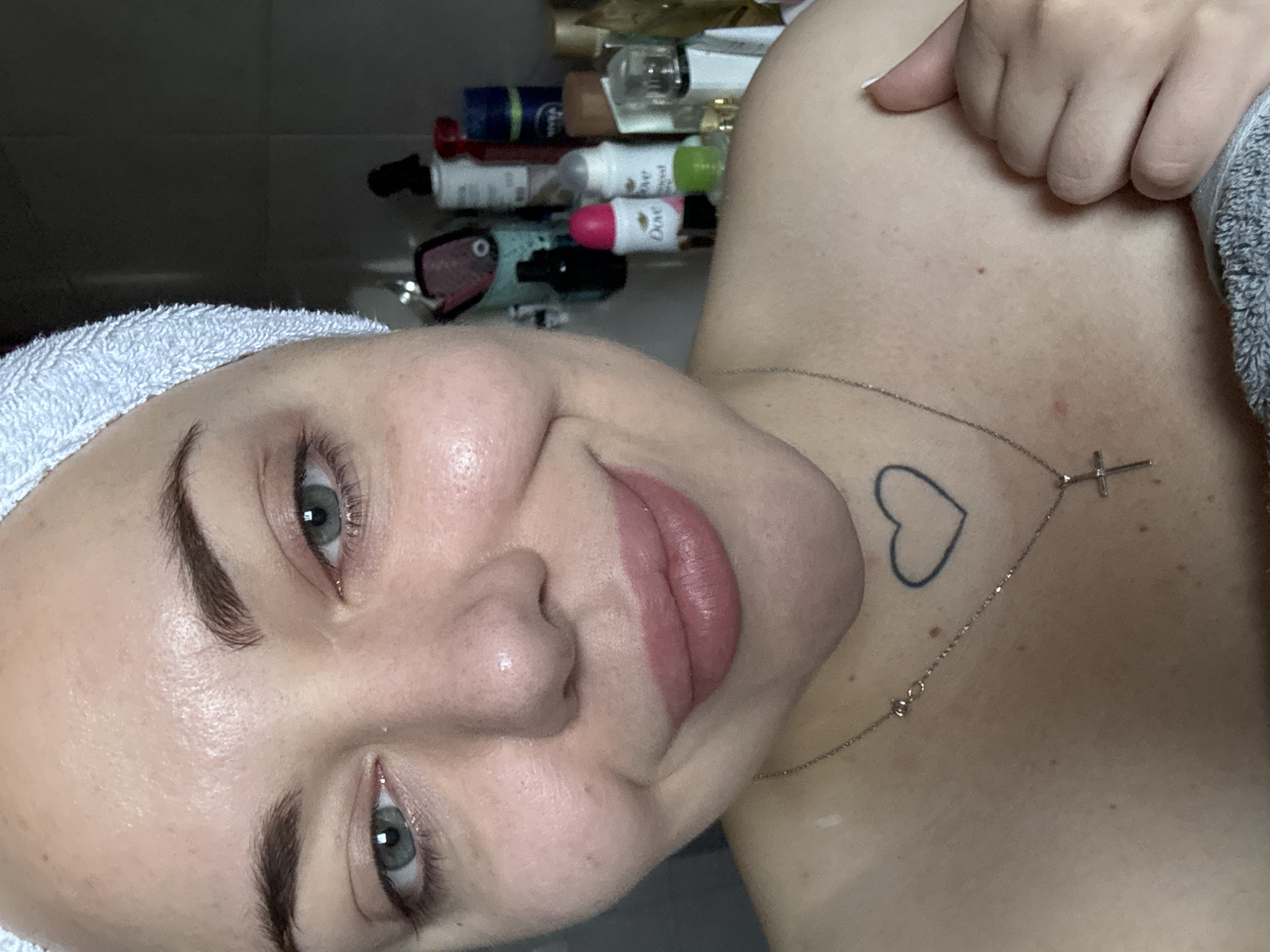 A close-up selfie of a woman wearing a white towel on her head and a necklace with a cross and a heart pendant. She has blue eyes and is wearing pink lipstick. Her skin appears smooth and clear. In the background, there are several bottles of toiletries on a shelf, some of which are partially visible, including Dove products. The scene is indoors, likely a bathroom, and the lighting is soft. There is no indication of the specific location within the city of Kremenchuk, Ukraine.