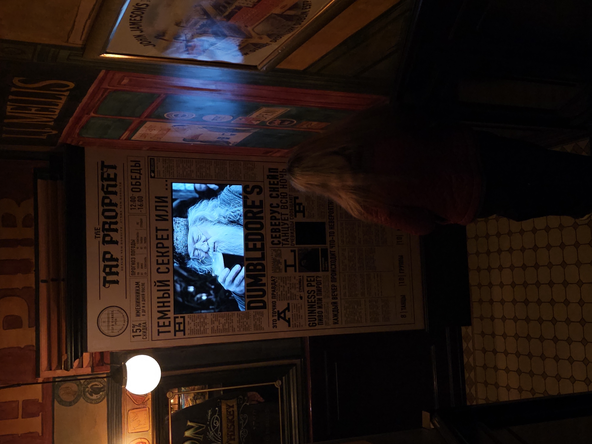 In an interior, dimly lit space, a woman stands facing a large, newspaper-style poster with a grainy image of Albus Dumbledore drinking from a goblet. The poster, titled "TAP PROPHET," features Russian text and headlines about a "Dark Secret." To the left of the poster is a glowing white spherical light fixture hanging from the ceiling. Further left, a dark sign with "ON WHISKEY" is visible. To the right of the poster, a dark green doorframe with red trim surrounds a display of what appears to be advertisements. Above the doorframe, the yellow lettering "LL MELLIS" is prominent. The floor is tiled in a diamond pattern of light and dark squares. The overall ambiance suggests a themed establishment.