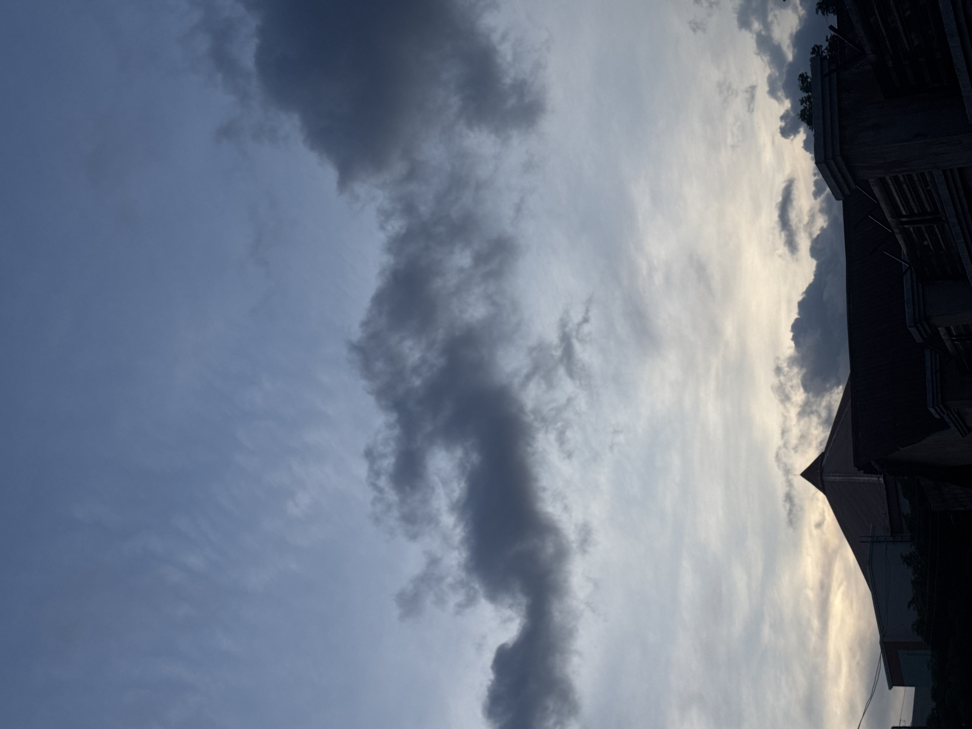 This is a full-frame, eye-level, vertical shot of a cloudy sky over buildings at dusk. The sky is predominantly blue with scattered dark clouds. The clouds are wispy and streaky in the upper portion of the image, becoming thicker and darker towards the right side. Below the sky, the silhouettes of buildings are visible. The most prominent building has a distinctive conical roof. Some structures appear to be residential or commercial. Trees and fencing are also visible in the lower portion of the image, adding to the urban landscape. The overall lighting suggests late evening or early morning, with the sun’s light diffused by the clouds. No people are visible in the image.