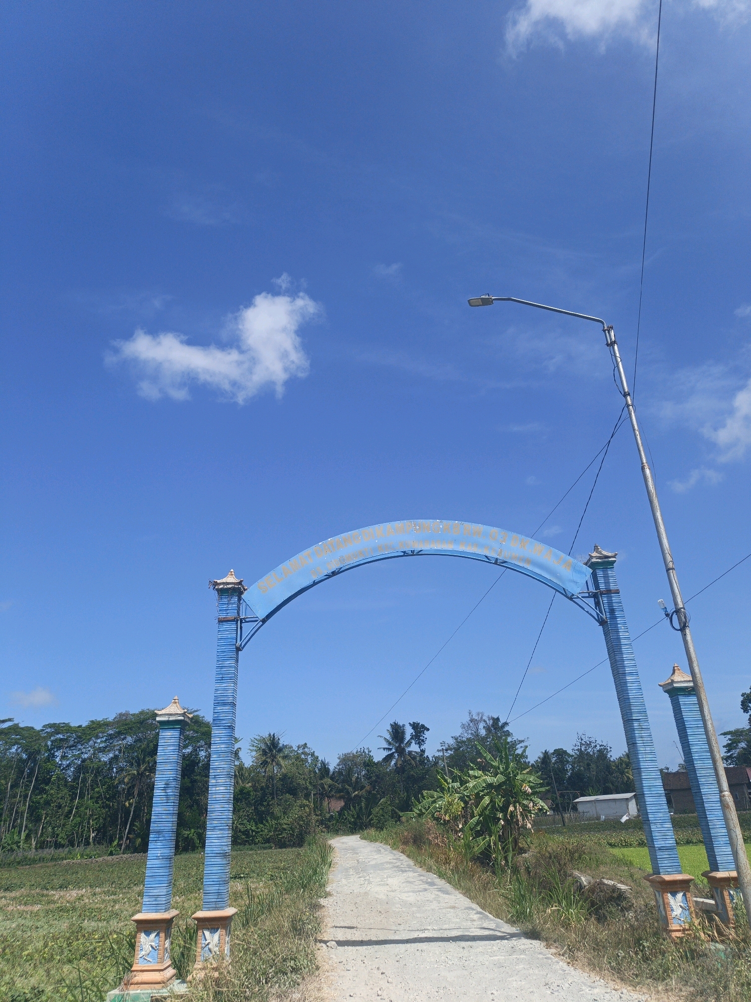 This is a low-angle, full outdoor shot of a rural landscape under a bright blue sky with scattered white clouds. The foreground features a paved path leading through overgrown grass and vegetation. On either side of the path, there are decorative blue and orange gate posts, with a blue archway spanning the path above. The archway has text written on it in gold, appearing to be a welcome or directional sign. Beyond the archway, the path continues into the distance, flanked by fields and lush green trees. A street lamp on a metal pole is visible on the right side of the image, with electrical wires extending from it. The environment suggests a village or agricultural area, possibly in the city of Kamulyan, Indonesia, given the architectural style and vegetation. There are no people or animals visible in the image.