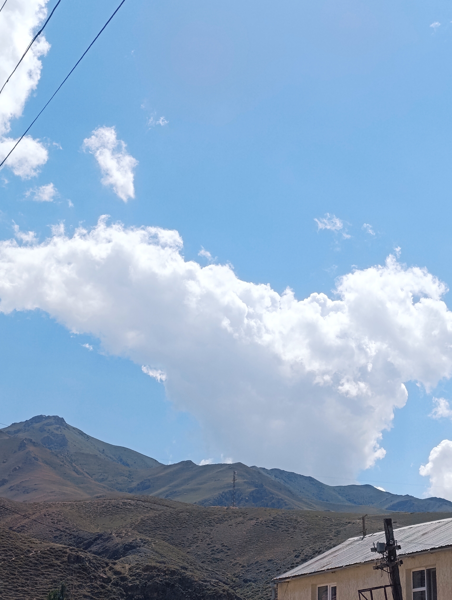 A daytime, eye-level, medium shot captures a wide view of rolling hills and mountains under a bright blue sky with scattered clouds. In the foreground, the roof of a light-colored building with a metal roof is visible, partially obscured by a utility pole with electrical equipment. The middle ground shows the textured, dry surface of lower hills, and in the background, the imposing, rugged peaks of larger mountains dominate the landscape. Overhead, thin, dark wires stretch across the upper portion of the frame. This scene is located in Naryn City, Kyrgyzstan.