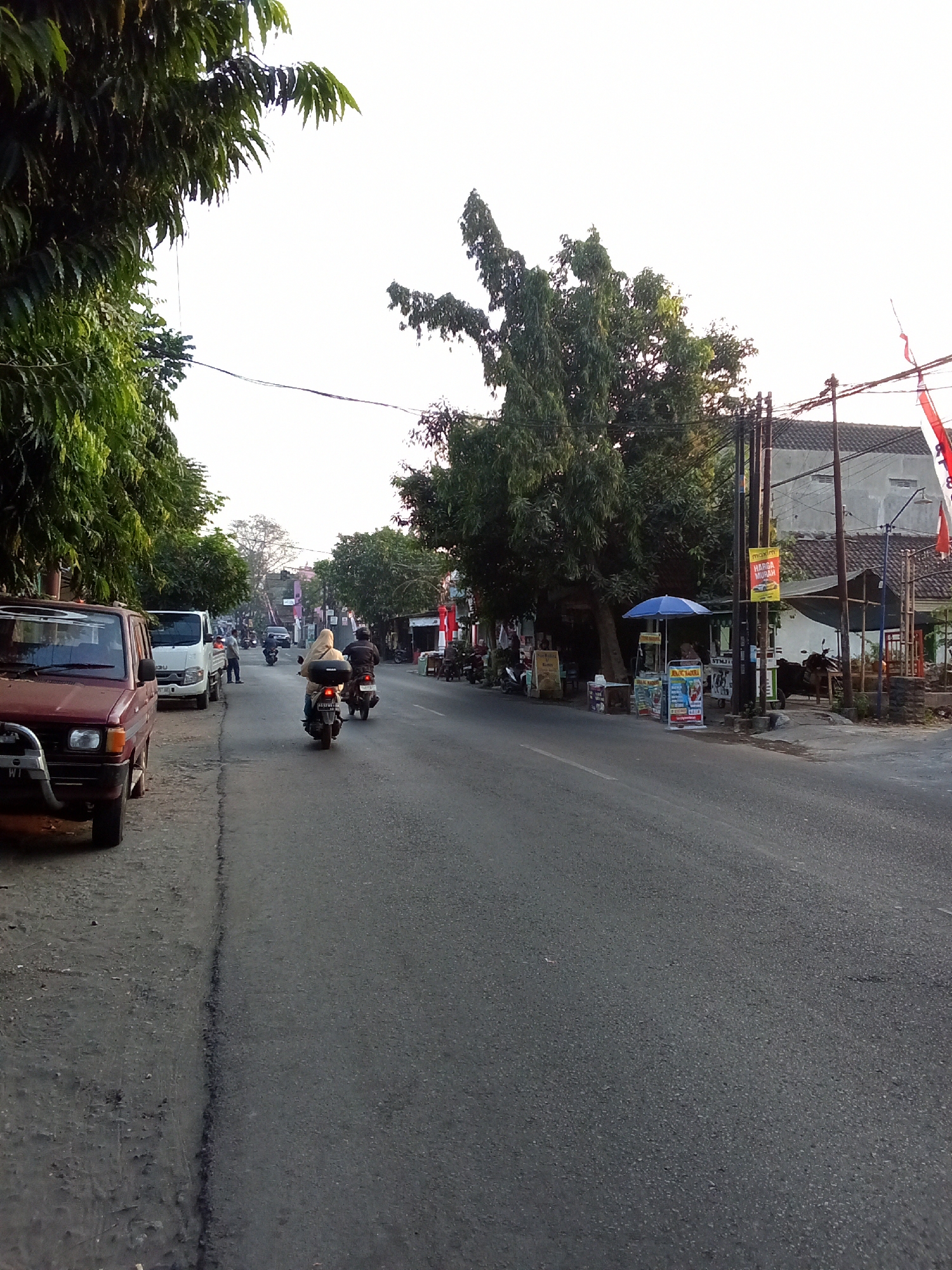 A street scene in Kediri City, Indonesia, is depicted. The image shows a paved road with a dirt shoulder on the left, lined with trees. Several vehicles are present, including a red car parked on the shoulder, a white van, and motorcycles on the road. People are seen riding motorcycles and standing near the parked van. On the right side of the road, there are small shops or stalls with umbrellas and signage. The sky is bright and appears to be daytime, possibly late afternoon due to the lighting. Buildings and utility poles are visible along the street.