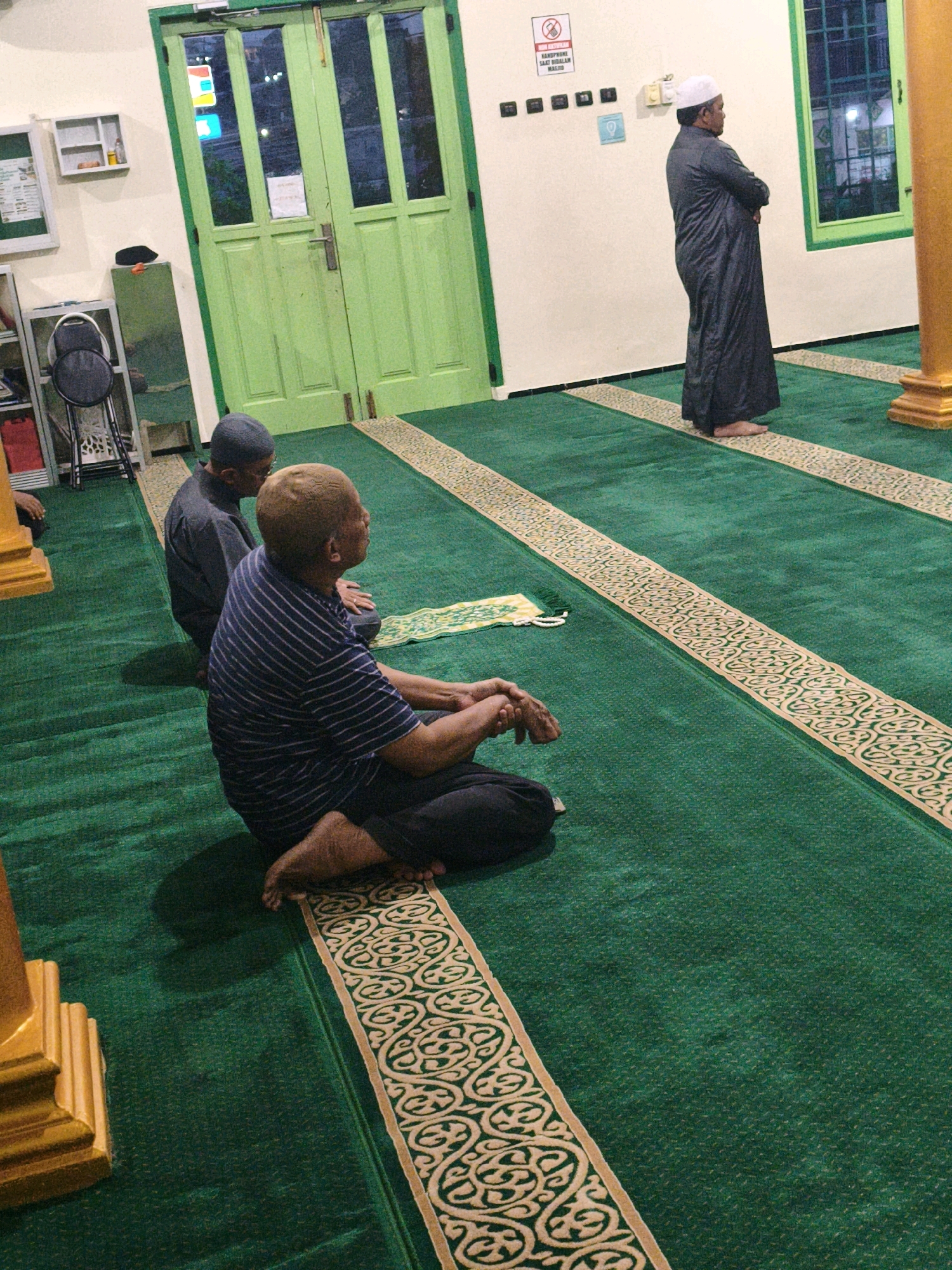 This image depicts the interior of a mosque in Samarinda, Indonesia. The flooring is covered with a vibrant green carpet featuring ornate, gold-colored borders. In the foreground, a person sits on the carpet, facing away from the camera, wearing a striped blue and white short-sleeved shirt and dark pants. Their legs are crossed, and their hands are clasped in their lap. To their left and slightly behind, another person is also seated on the carpet, facing the same direction, wearing dark clothing and a dark cap. Further back, a third individual stands, facing away from the camera and towards a wall, wearing a dark robe and a white cap. This person's arms are crossed. The background reveals a green door with multiple glass panels, allowing a glimpse of the exterior. To the left of the door, shelves are mounted on the wall, one containing what appears to be a red object. A metal chair is also visible. On the wall, there are signs, including one with a red circle and diagonal line, indicating a prohibition, and another with text that is partially obscured. The overall impression is a serene and respectful atmosphere within the mosque.