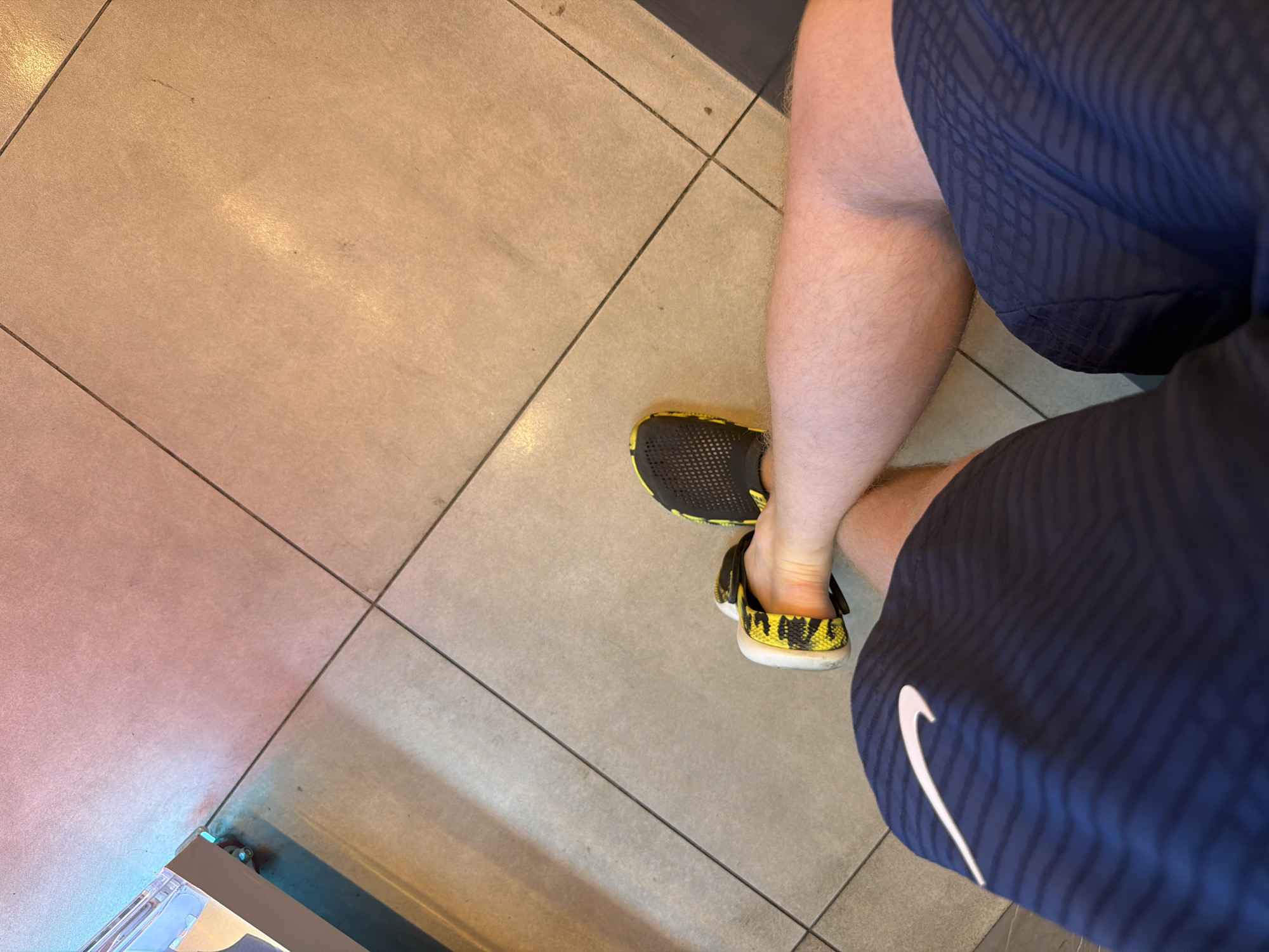 A person's legs are shown from a top-down perspective. They are wearing navy blue shorts with a white Nike logo and dark yellow and black patterned athletic shoes. The person is seated on a light brown tiled floor, which is intersected by dark grout lines. The lighting in the scene is warm and appears to be artificial, casting shadows. The background shows a wall with a light colored baseboard and some blue light reflecting from an unseen source. Based on the available visual information, it is not possible to determine the specific location within the city of Kyiv, Ukraine, or the time of day.