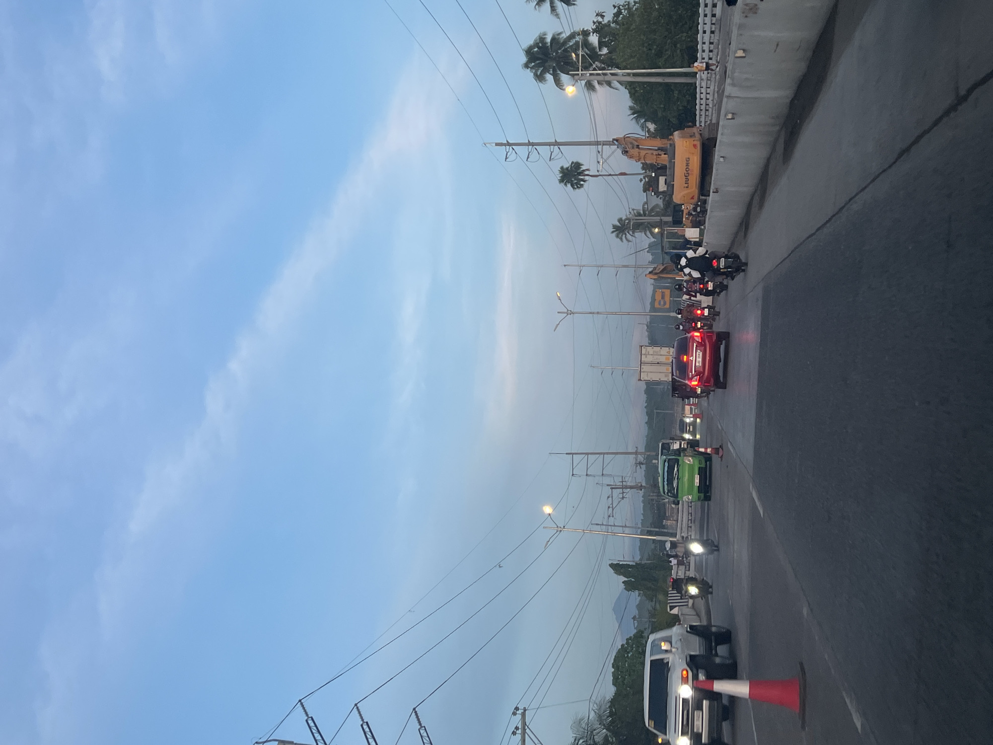 A traffic scene unfolds on a highway in Davao City, Philippines, under a light blue sky with scattered white clouds. The time appears to be either early morning or late afternoon, given the ambient lighting. A white SUV is visible on the left side of the road, with its headlights on. A red traffic cone is placed in front of it. Further down the road, several other vehicles, including a green van, motorcycles, and cars, are moving or stopped. Construction equipment, specifically an excavator with the brand "LIUGONG" visible, is present on the right side of the highway, indicating ongoing road work. Palm trees and other foliage line the sides of the highway, and power lines crisscross the sky. In the background, a distant conical mountain shape is faintly visible, a characteristic landmark of Davao City, likely Mount Apo. People are seen on motorcycles and walking near the construction area. The road surface is asphalt, marked with white lines.
